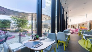 Un área de comedor exterior moderna con mesas y sillas, ventanas amplias y vistas al paisaje urbano.