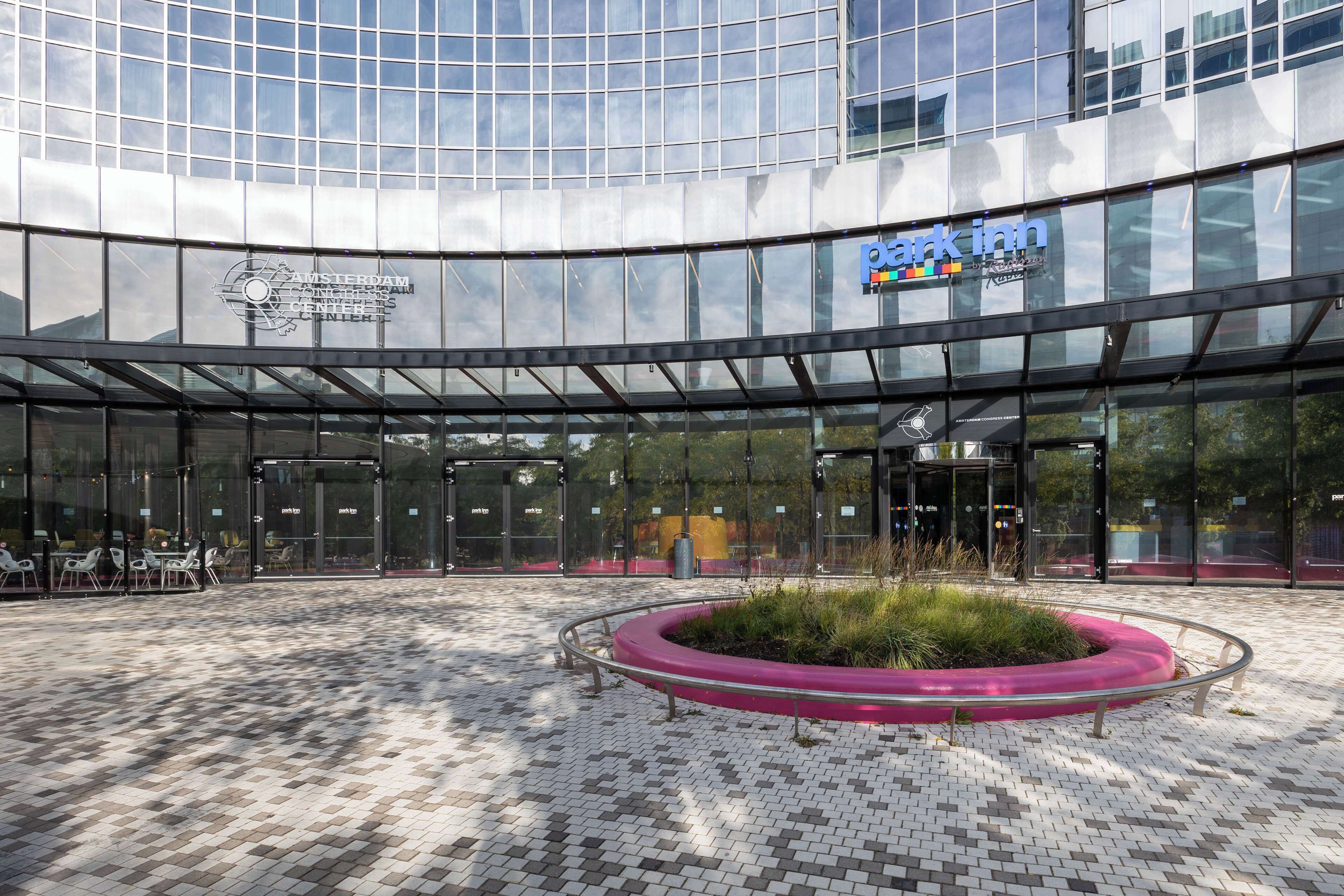 Un bâtiment moderne à la façade vitrée, avec un grand jardinier circulaire planté de verdure au centre, et une enseigne « Park Inn » sur la droite.