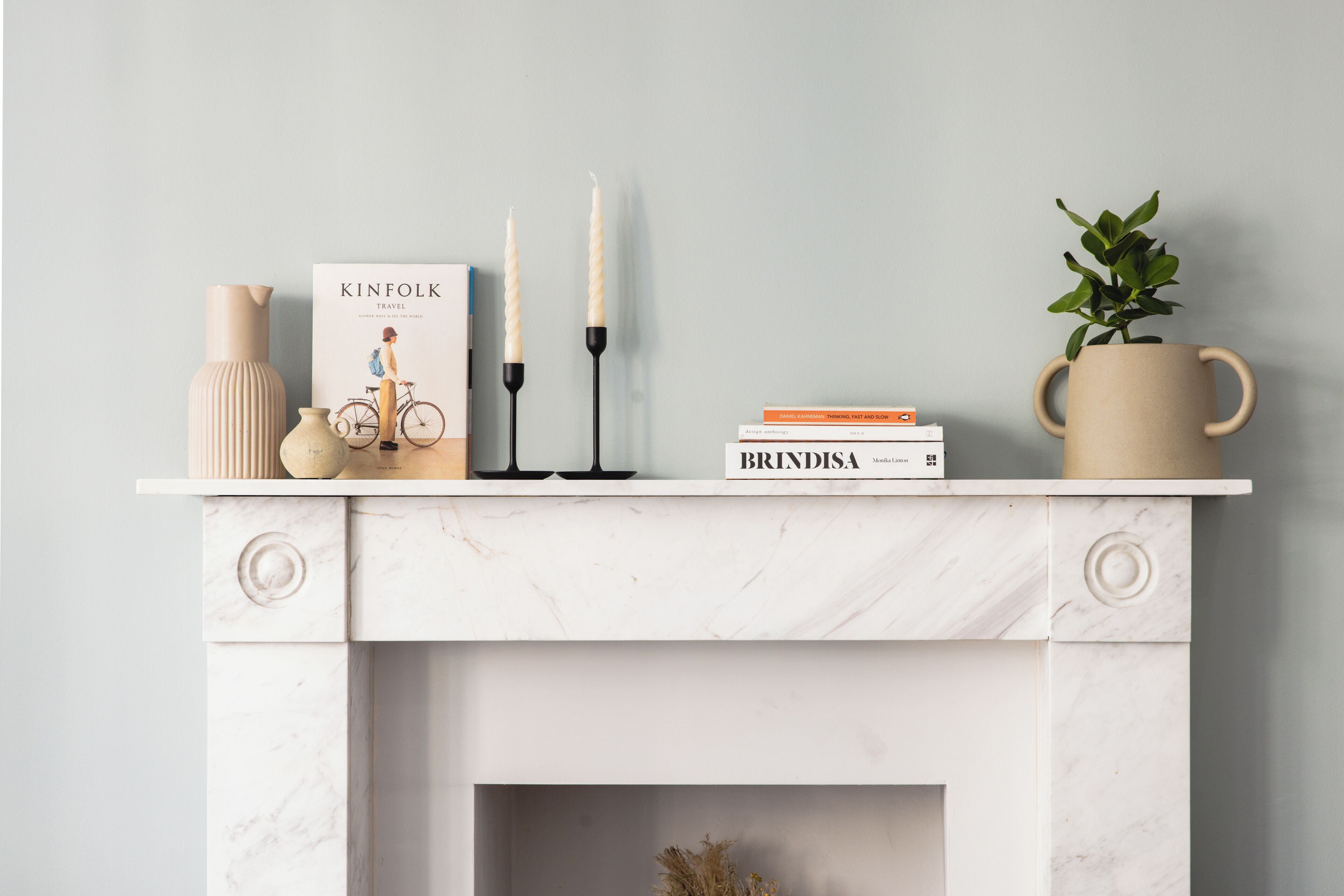 A fireplace mantel with a book titled 'KINFOLK TRAVEL,' two candles, a vase, a potted plant, and a mug.