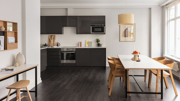 A modern kitchen with dark cabinets, a white countertop, and a dining area with a white table and wooden chairs. - Native Fenchurch  (London)
