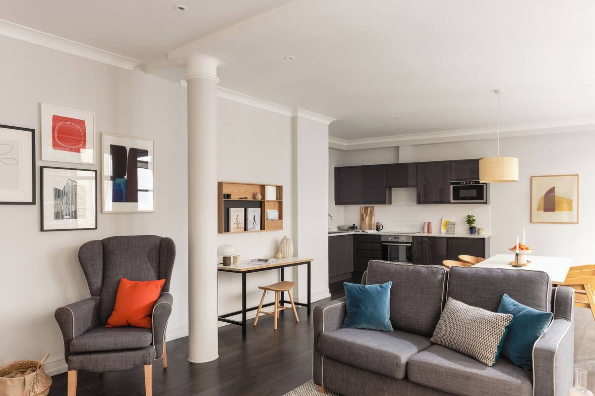 A modern living room with a grey sofa and armchair, a small wooden table, and a kitchen in the background.