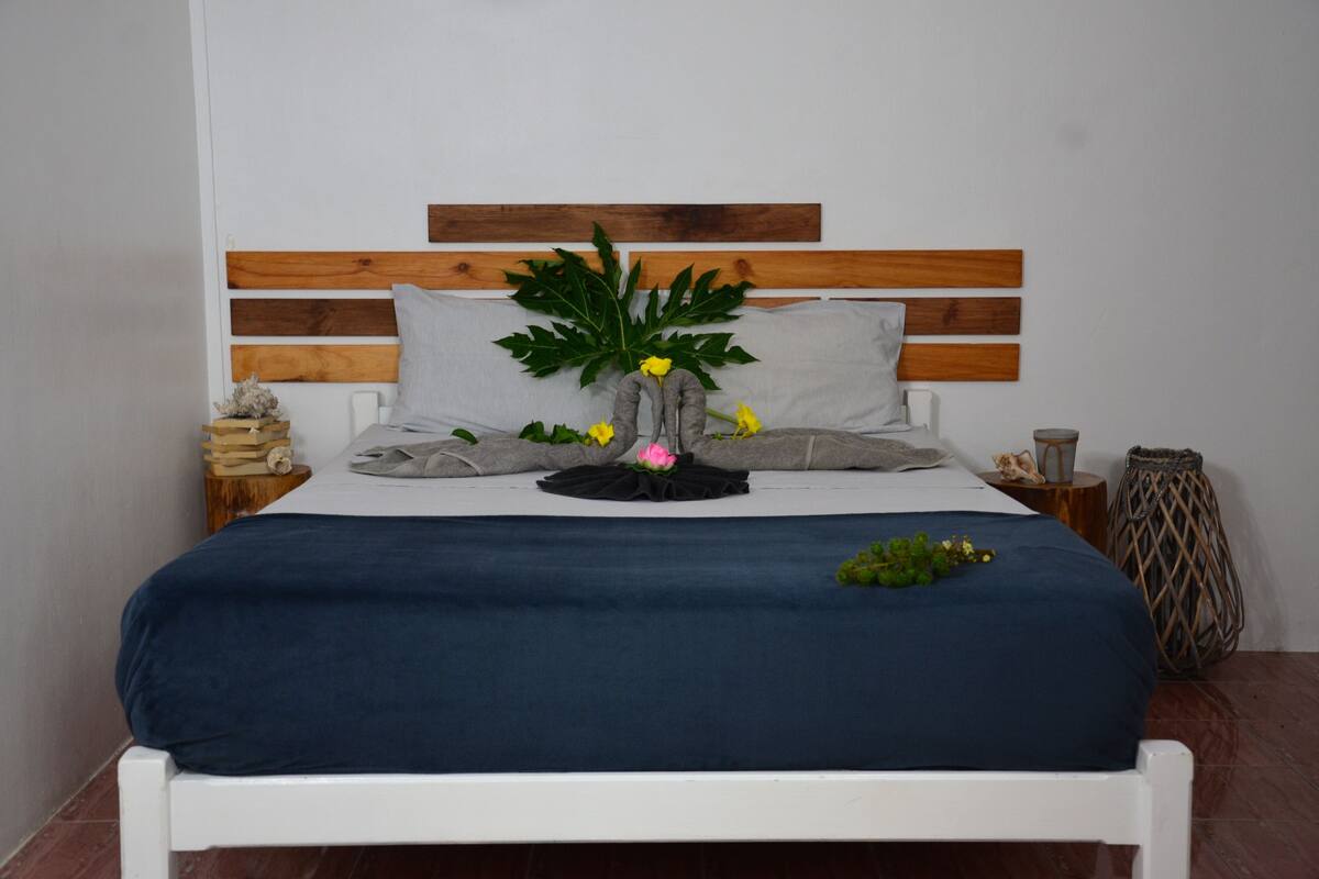 Ein Bett mit Holz-Kopfteil, grauen Kissen und einer blauen Tagesdecke.