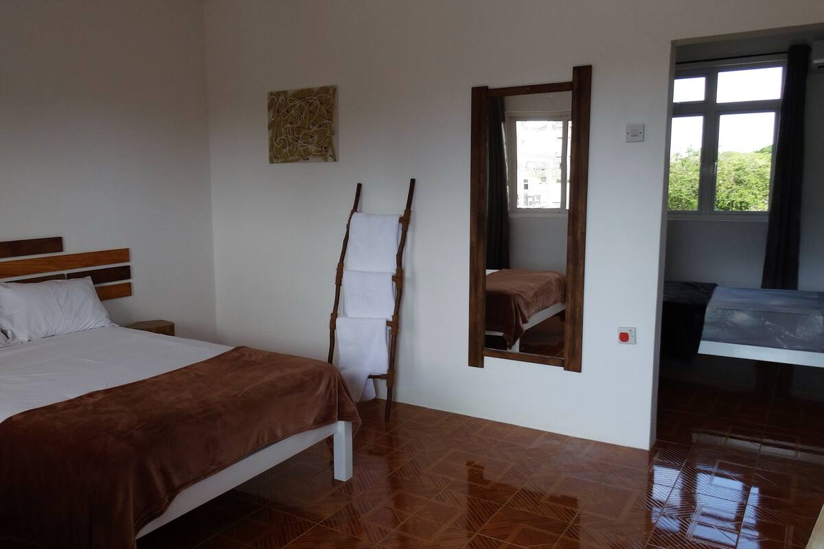 Ein Schlafzimmer mit einem Bett, einem Holzkopfende, einem an der Wand befestigten Spiegel und einem Fenster mit Blick auf Grünflächen.