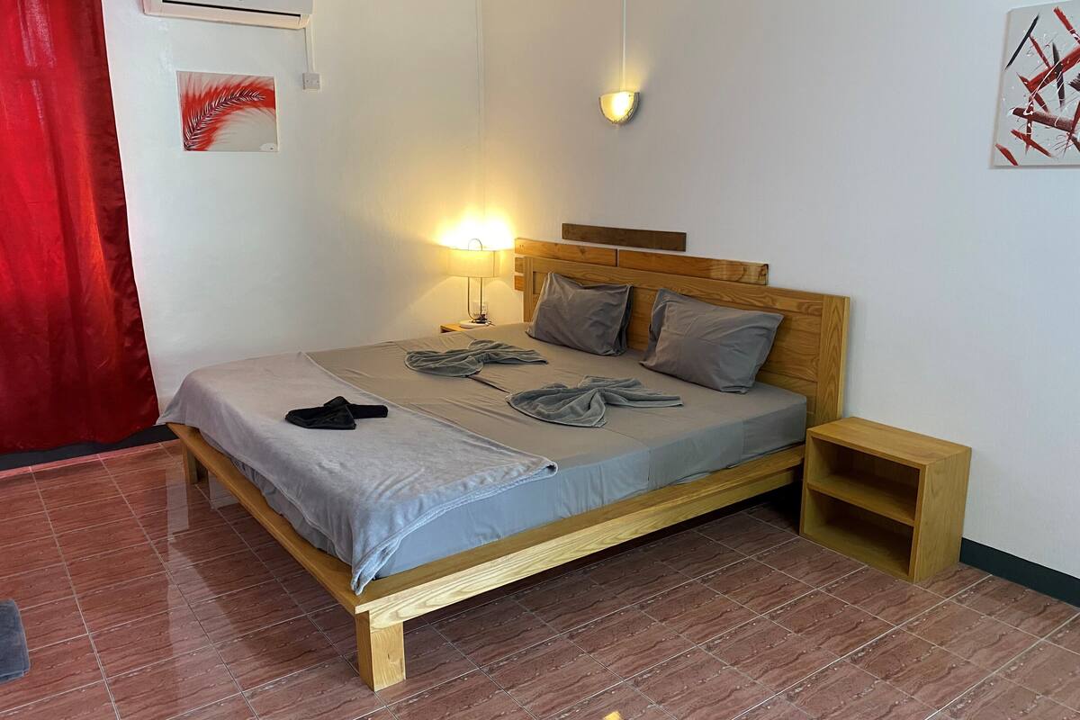 Ein ordentlich bezogenes Bett mit Kissen und Decke, ein Nachttisch, eine Wandlampe und eine Klimaanlage.