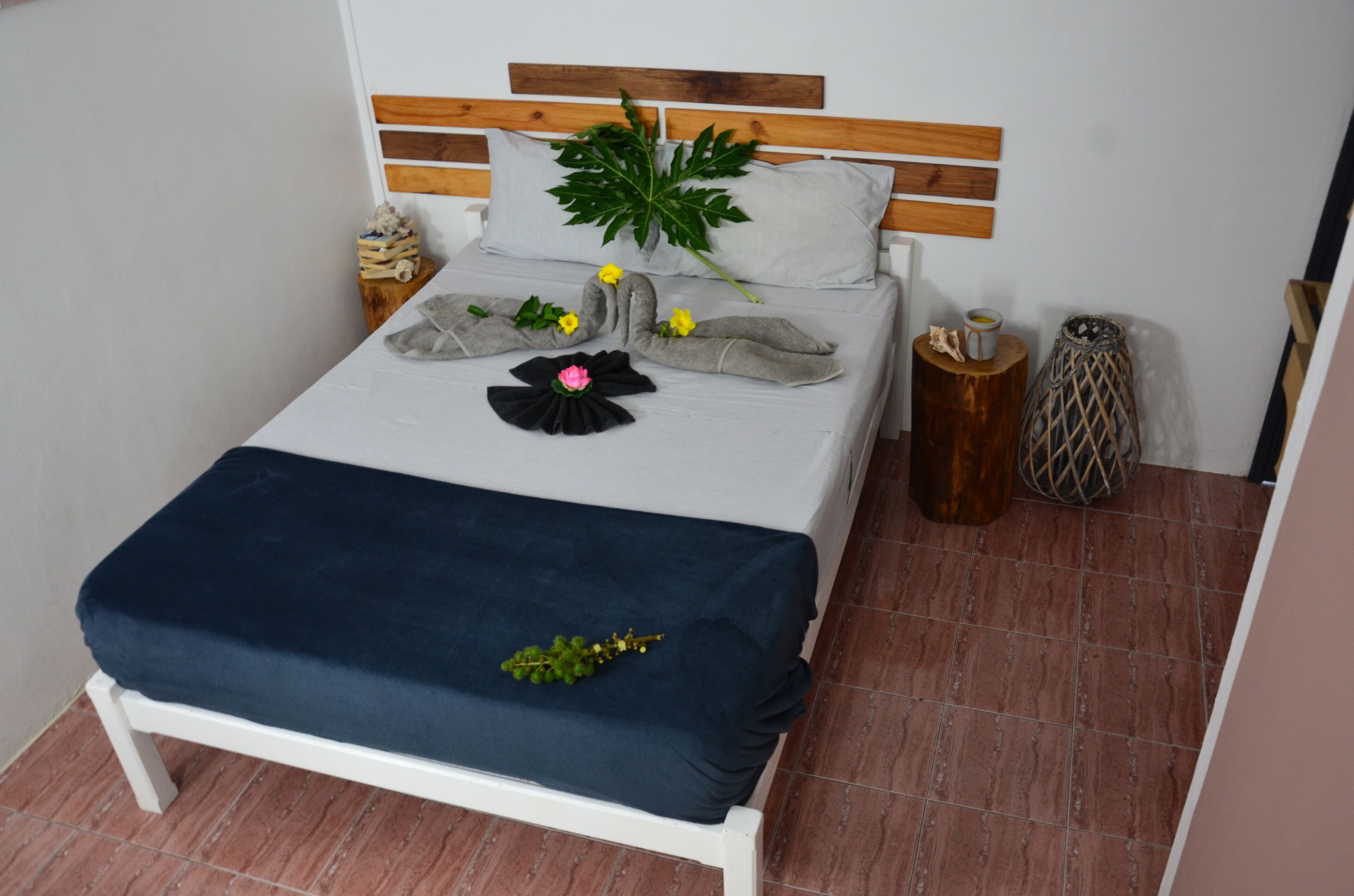 Ein ordentlich bezogenes Bett mit einer dunkelblauen Tagesdecke, einem Holzkopfstück und dekorativen Kissen, darunter ein großes grünes Blatt und eine schwarze Blume.