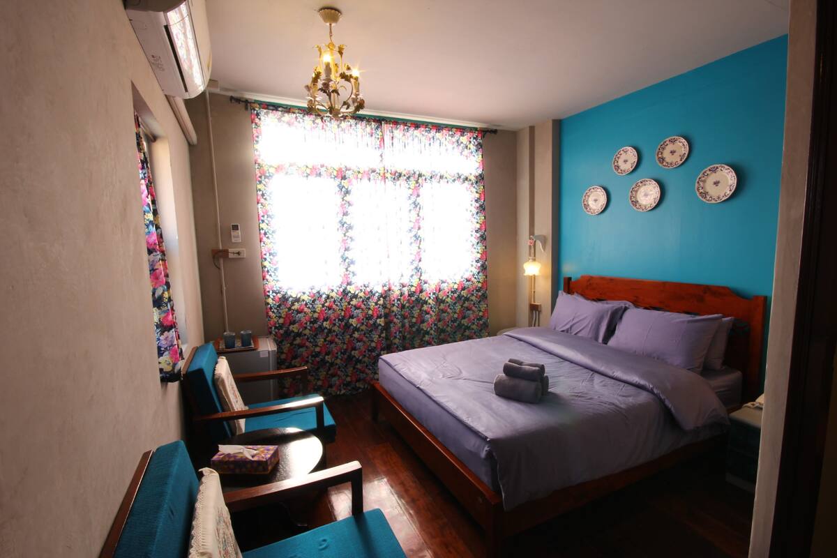 Ein Schlafzimmer mit Bett, Schreibtisch, Stuhl, einem Fenster mit blumigen Vorhängen und Wanddekorationen.