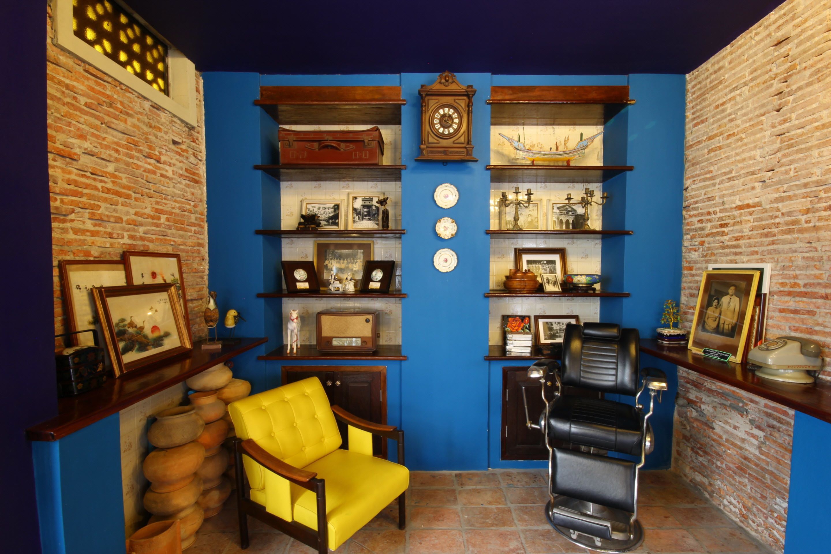 A room with a brick wall, blue painted walls, a yellow armchair, a barber chair, shelves with various items, and a clock.