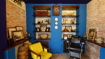 A room with a brick wall, blue painted walls, a yellow armchair, a barber chair, shelves with various items, and a clock.
