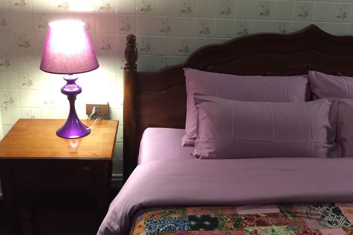 Ein Schlafzimmer mit Nachttisch aus Holz, einer violetten Lampe, einem Bett mit violetter Bettdecke und Kissen sowie einer Wand mit Blumenmuster.