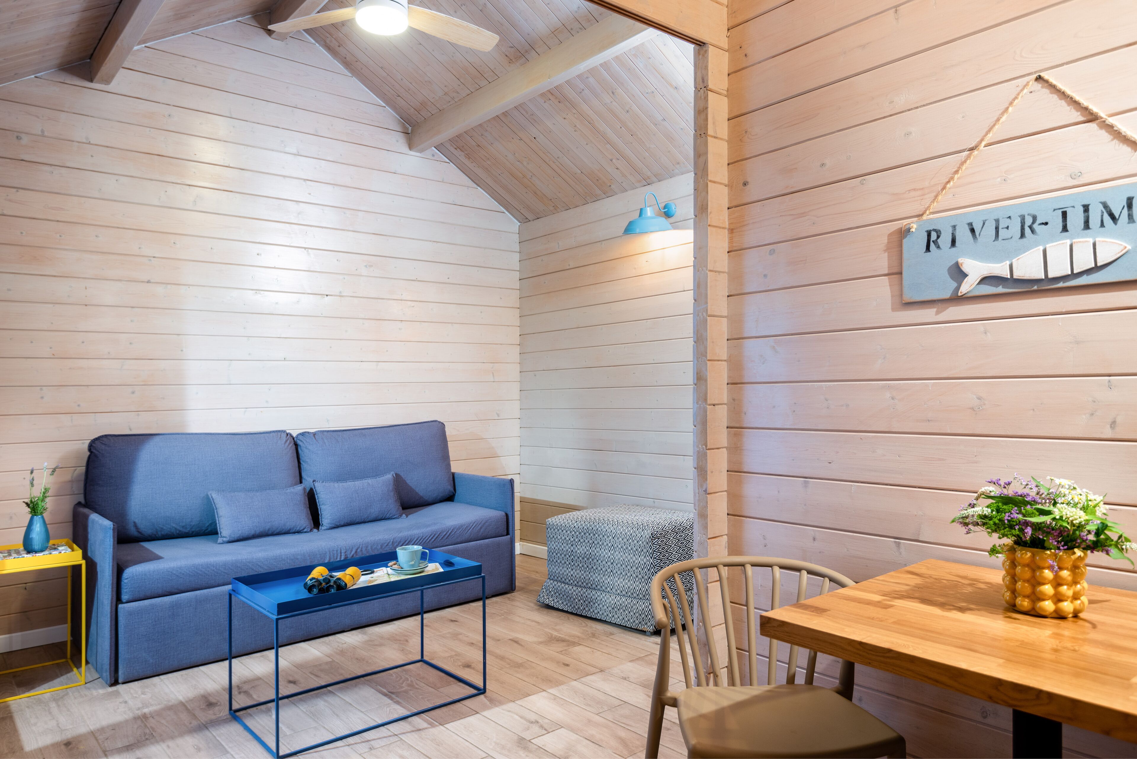 A cozy room with a blue sofa, a wooden table with a plant, and a sign reading "RIVER-TIME".