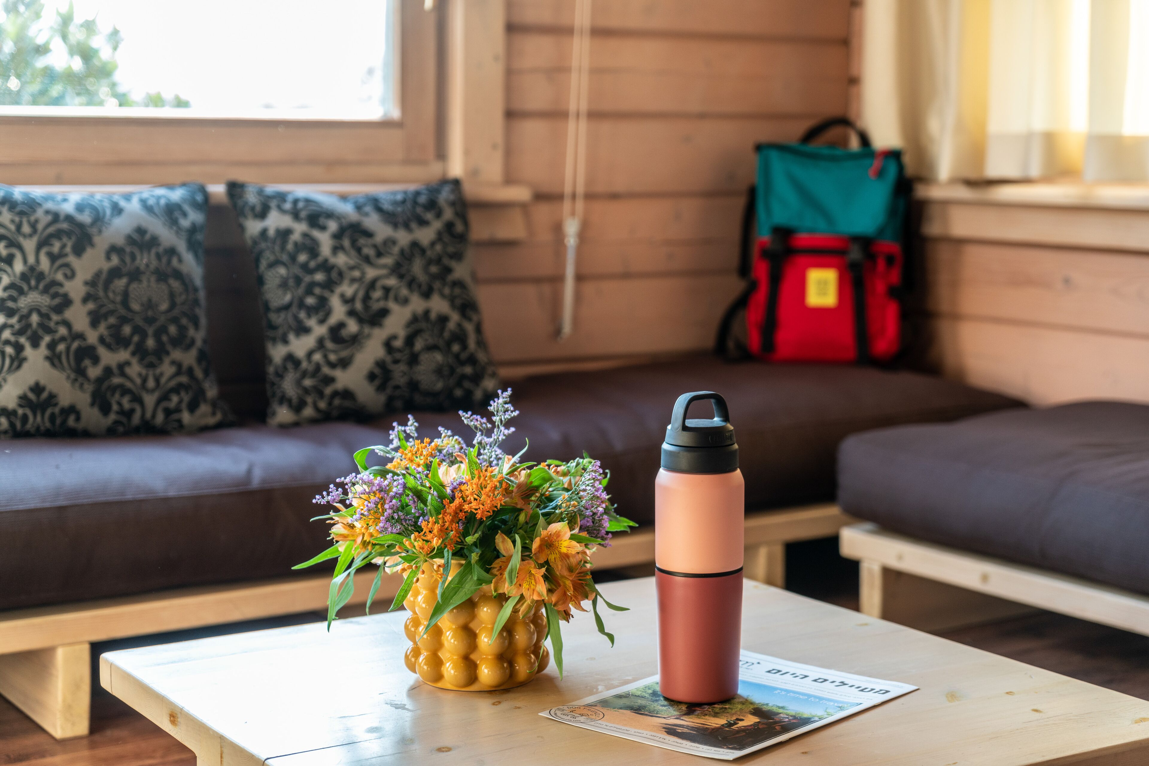 Una habitación acogedora con un sofá de madera, una mesa con un termo y un florero, y una ventana con vistas a árboles.