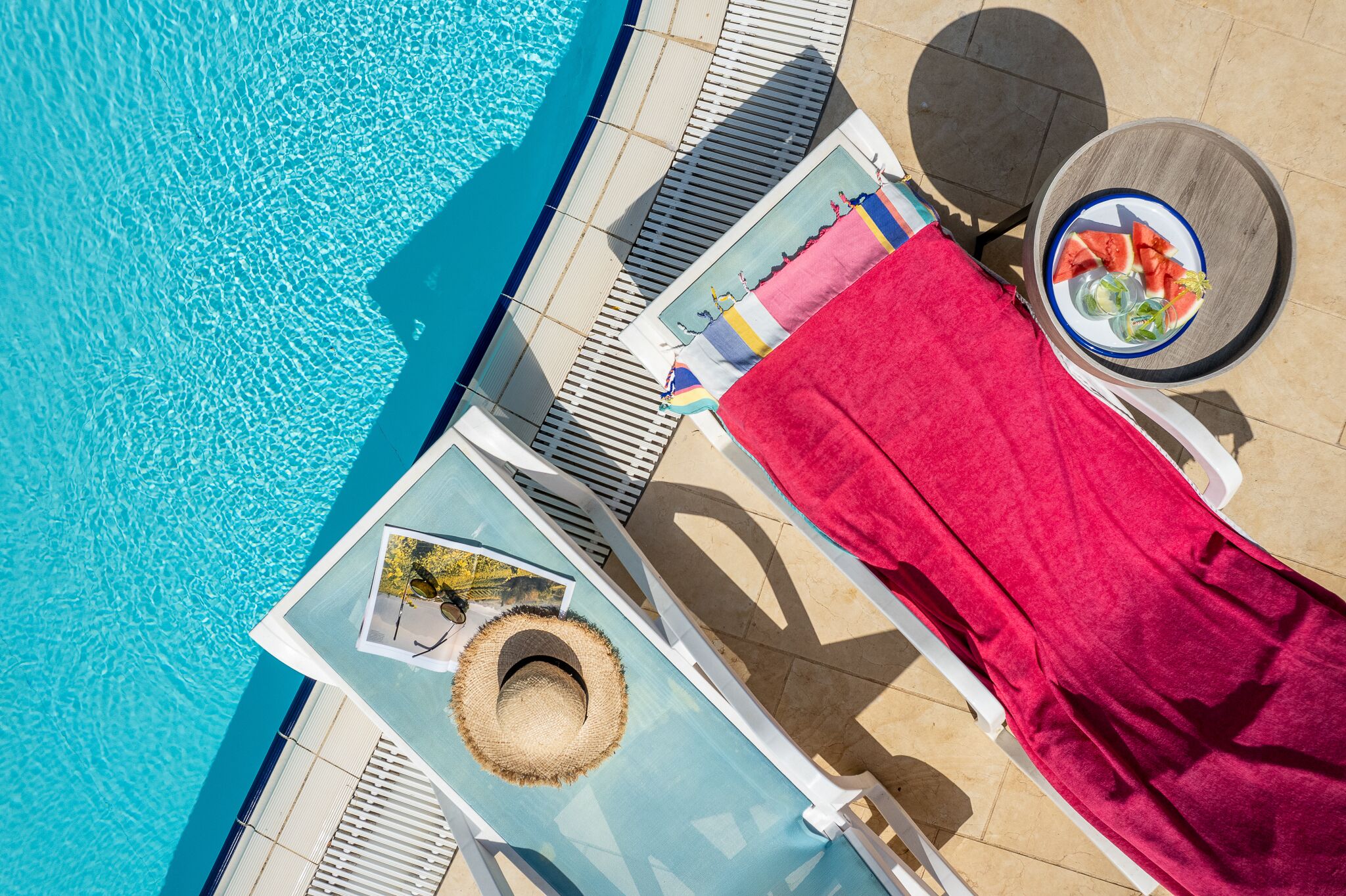 Zona de piscina con sillas de descanso, una mesa con sandía y menta, y una toalla roja.