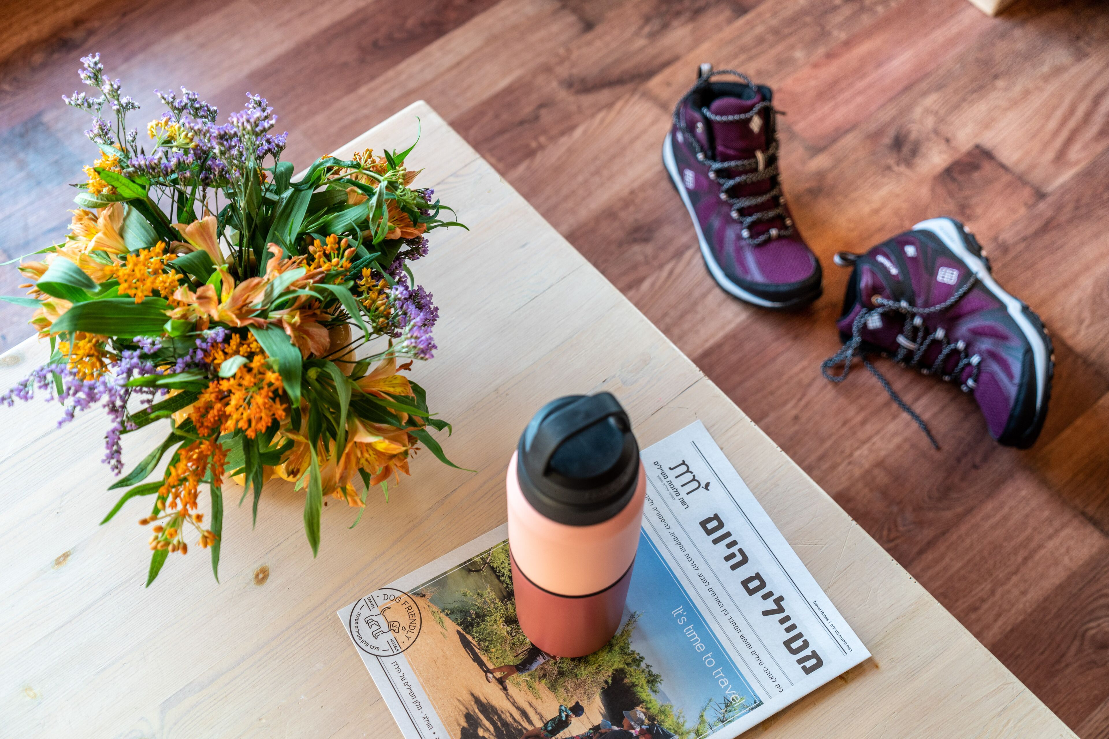 A wooden table with a flower arrangement, a thermos, a pair of hiking boots, and a magazine.