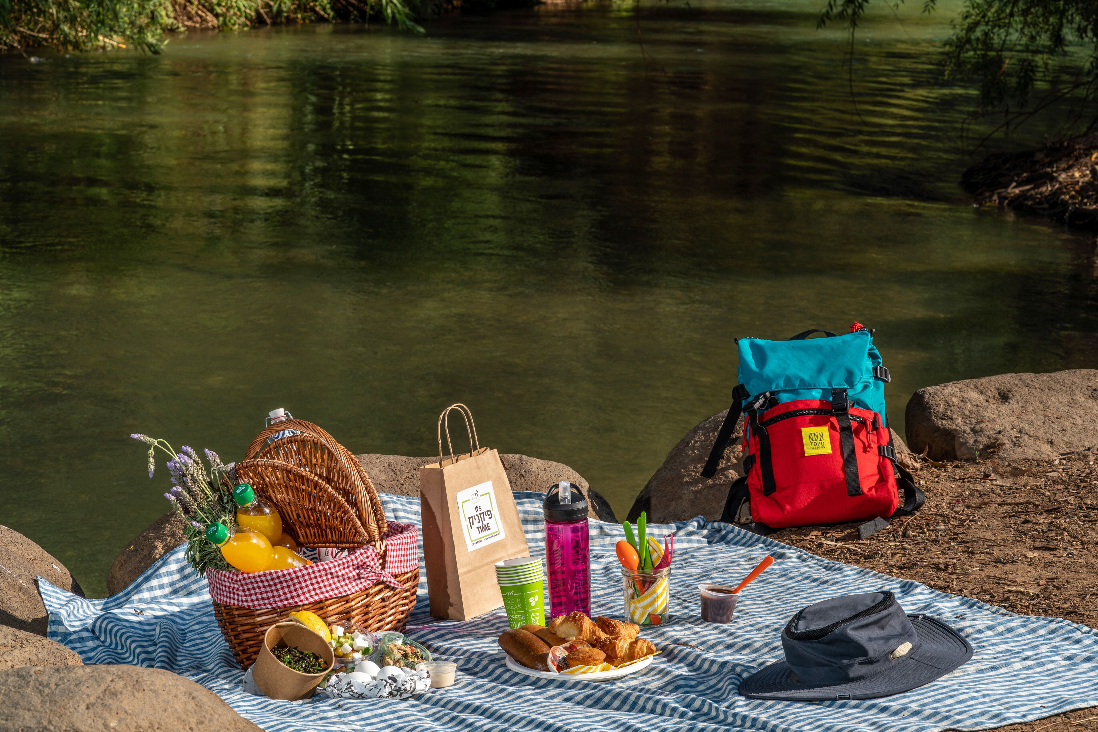 Un montaje para picnic junto a un río, con una cesta, comida, bebidas y una bolsa.