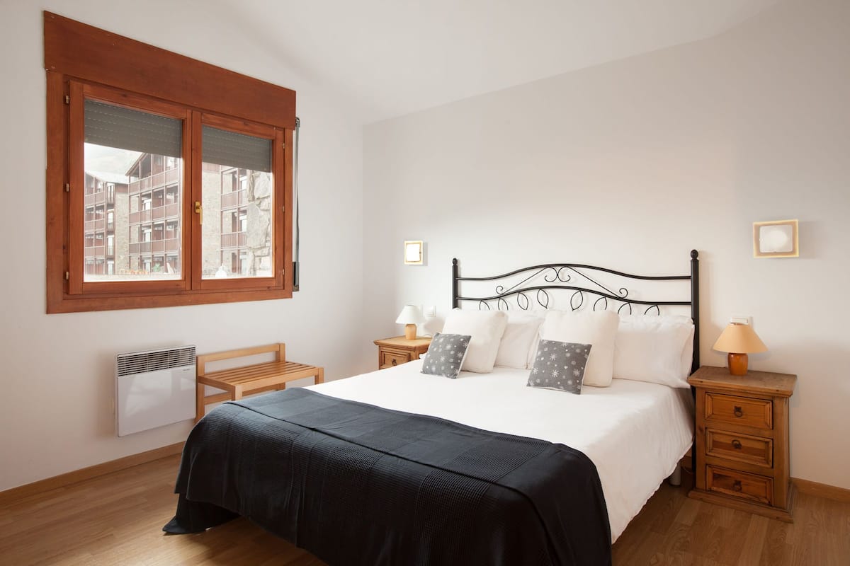 A bedroom with a bed, wooden nightstands, a window with blinds, and a radiator.