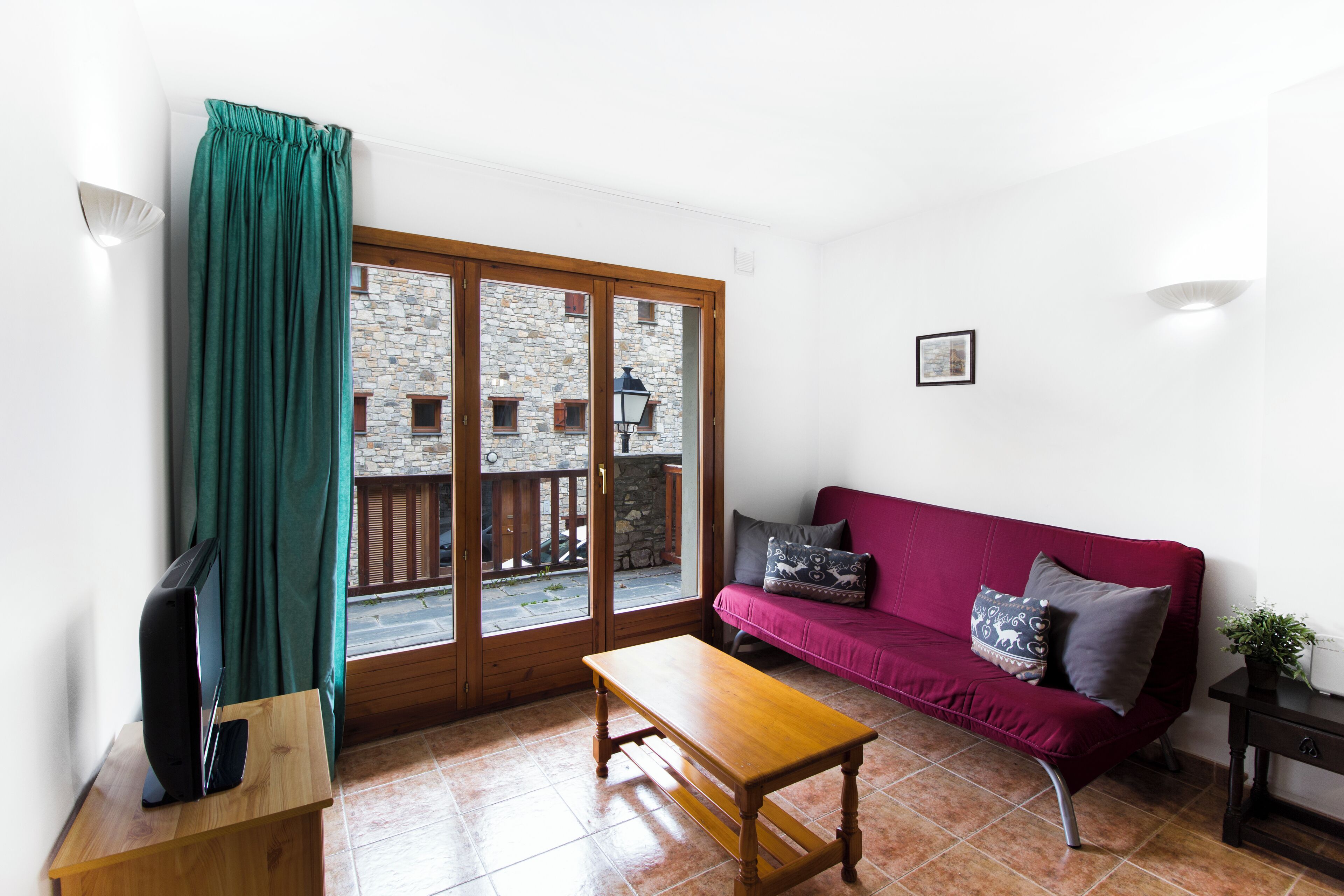 Una sala de estar con una mesa de madera, un sofá rojo con cojines estampados, un televisor sobre un mueble bajo y vista a un edificio de piedra a través de una puerta de cristal.