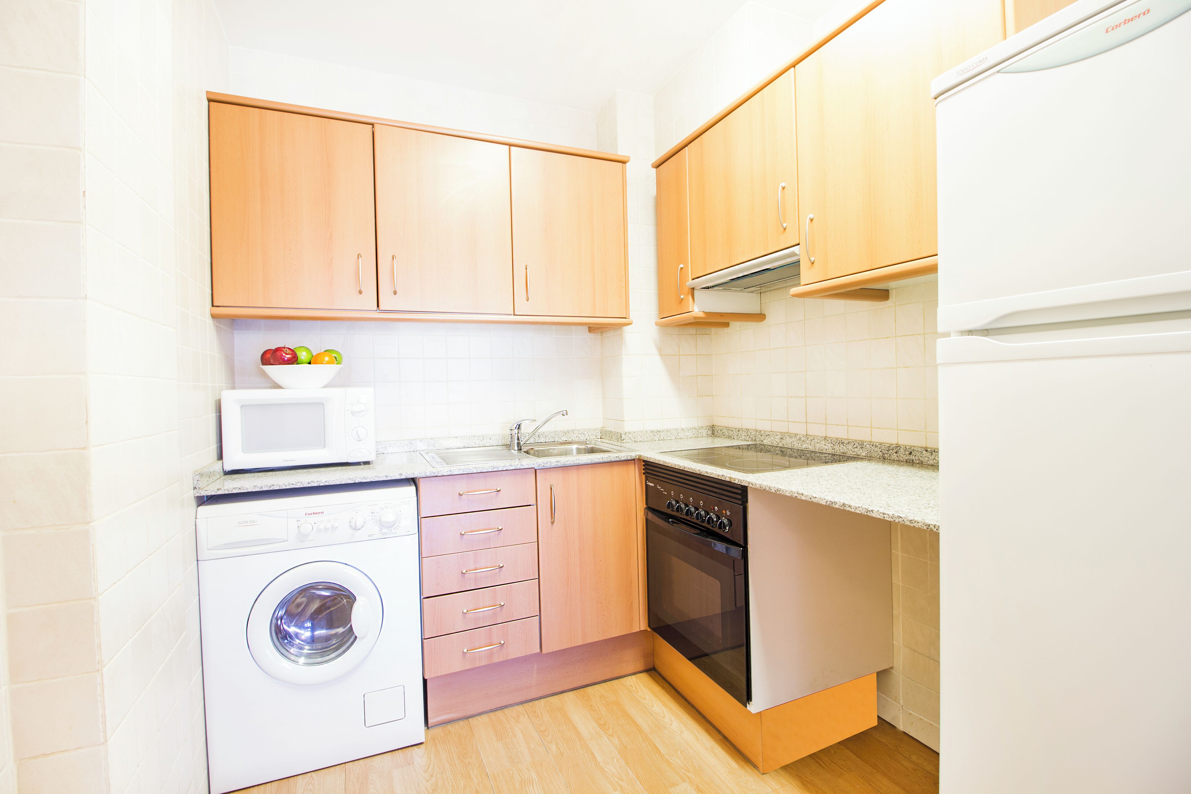A compact kitchen with wooden cabinets, a washing machine, a microwave, and a dishwasher.