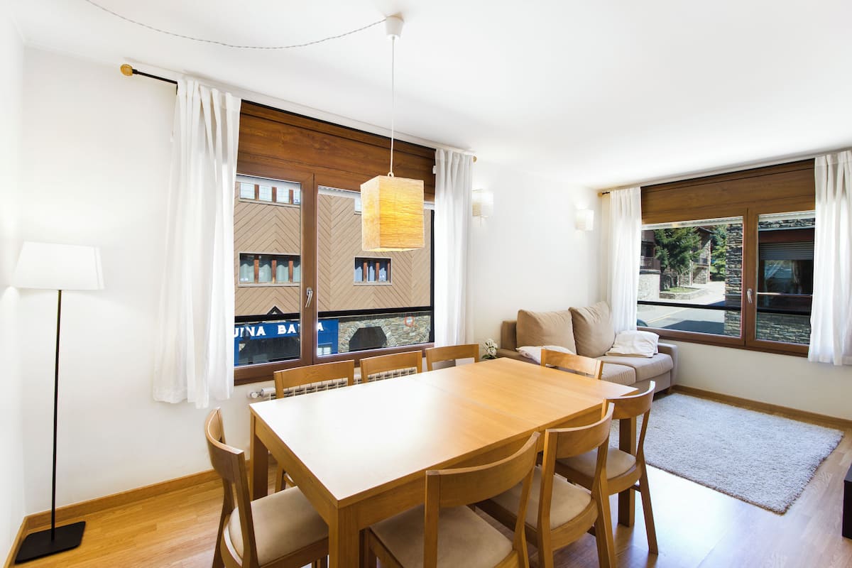 A modern dining area with a wooden table and chairs, a sofa, and large windows offering a view of the outside.