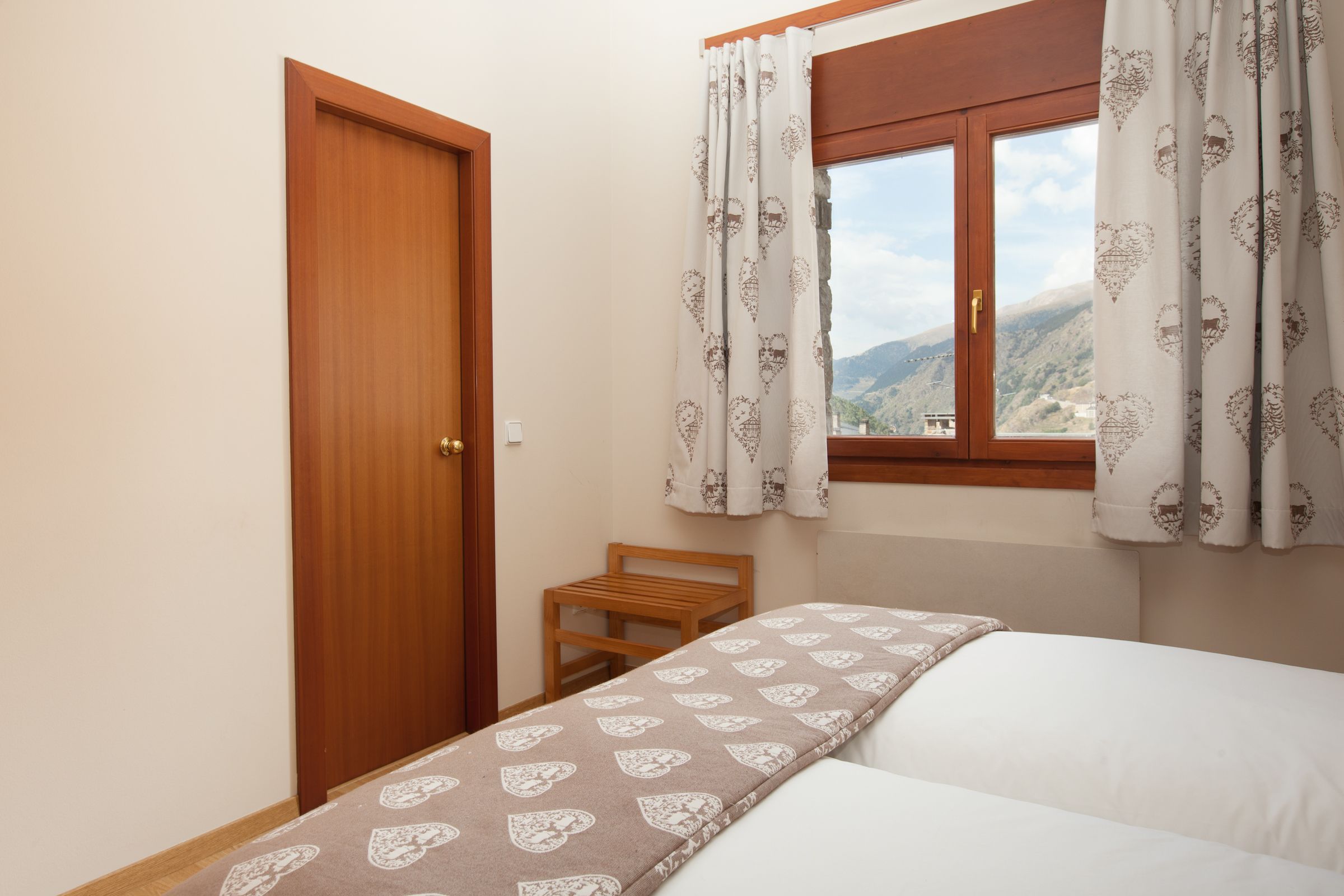 Une chambre à coucher avec un lit, une porte en bois, une fenêtre ornée de rideaux à motifs et une vue sur un paysage montagneux.