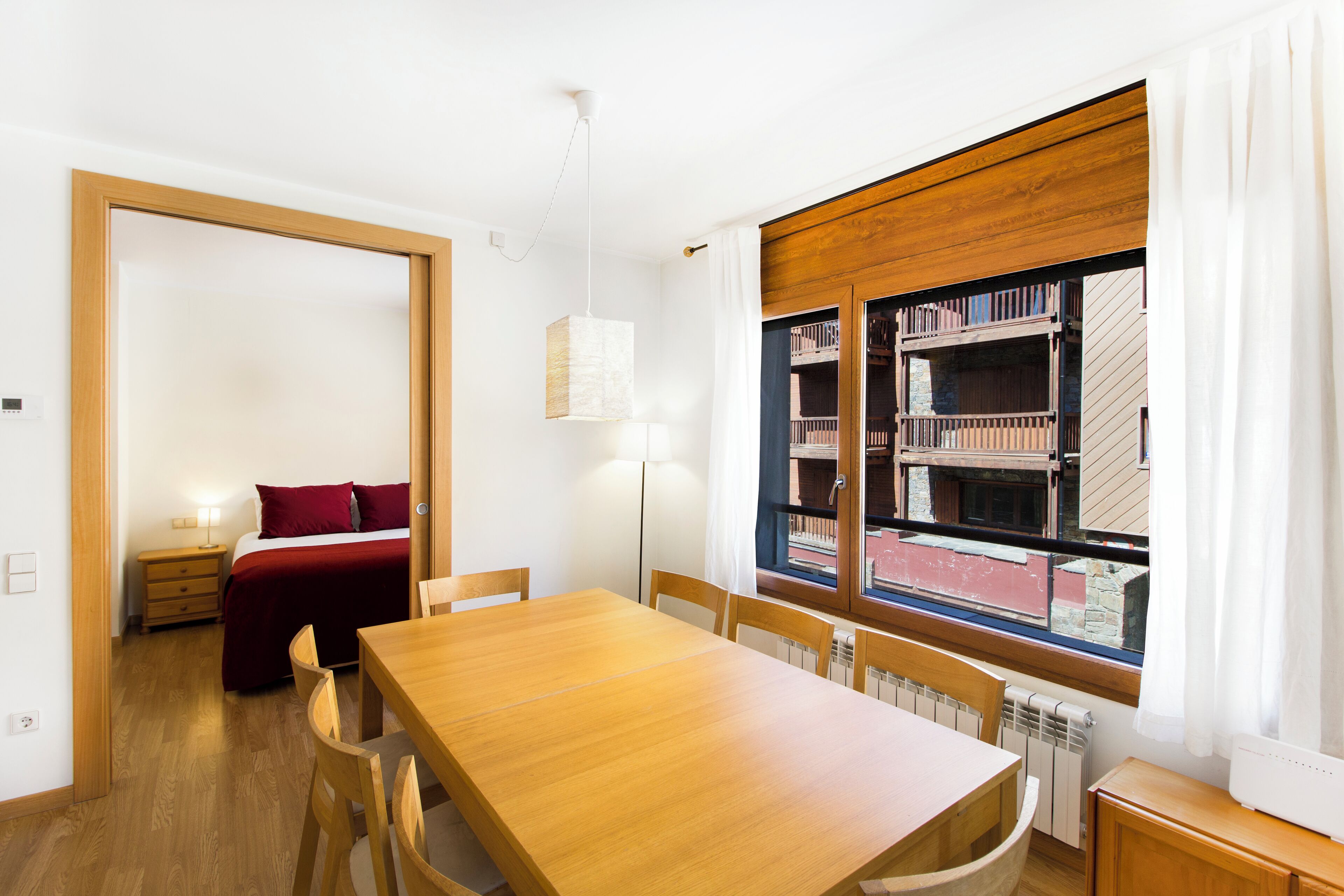 A room with a wooden dining table, chairs, a bed with red bedding, a bookshelf, and a window with curtains.