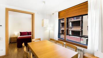 Una habitación con una mesa de comedor de madera, sillas, una cama con ropa de cama roja, una estantería y una ventana con cortinas.