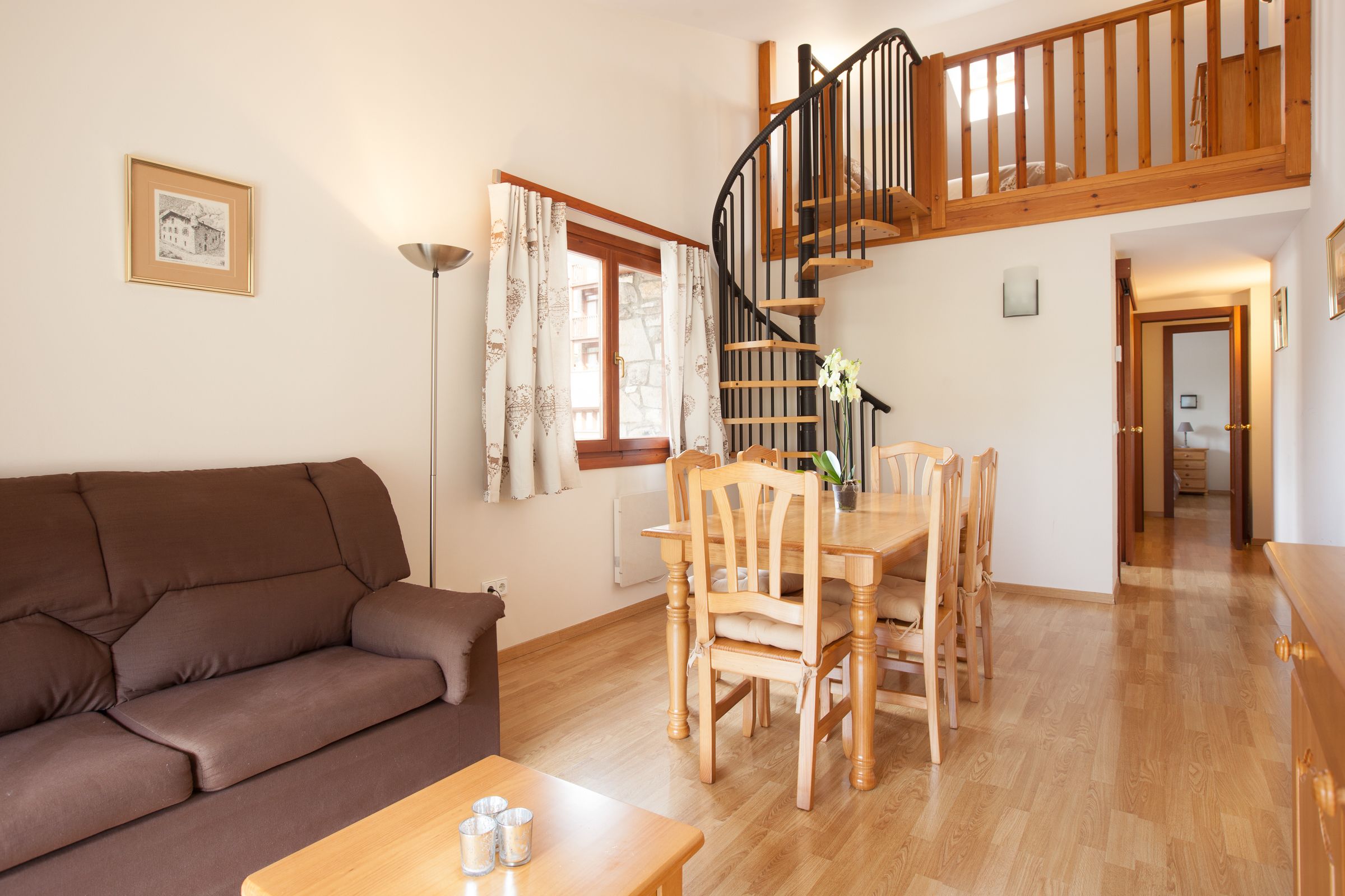 Una sala de estar con un sofá marrón, una mesa de comedor de madera y sillas. Una escalera con barandillas de madera conduce a un piso superior.