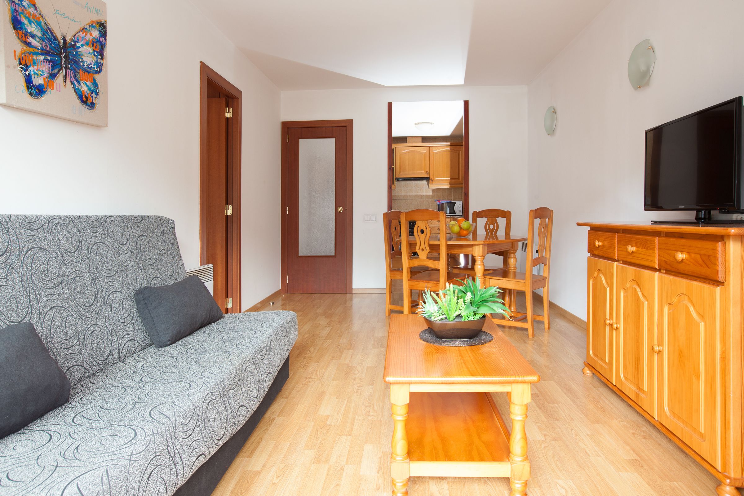 Ein Wohnzimmer mit einer Couch, einem Couchtisch mit Pflanze, einem Esstisch mit Stühlen, einer Küchenzeile und einem Fernseher auf einer Holzkommode.