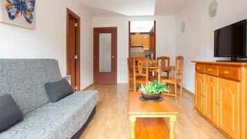 Una sala de estar con un sofá, una mesa de centro con una planta, una mesa de comedor con sillas, una zona de cocina y un televisor sobre un mueble de madera.