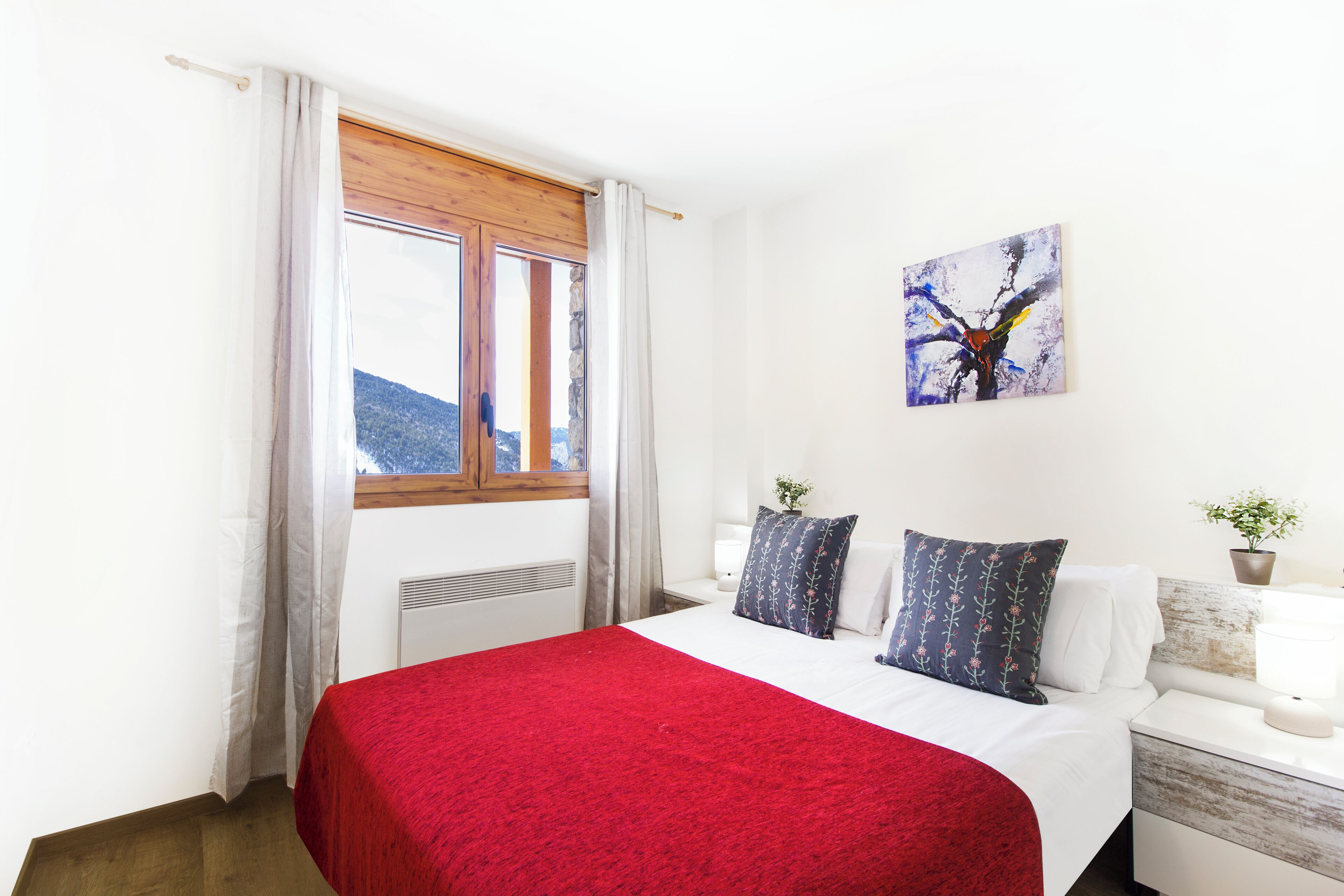 A bedroom with a bed, a window with curtains, a nightstand with a lamp, and a painting on the wall.