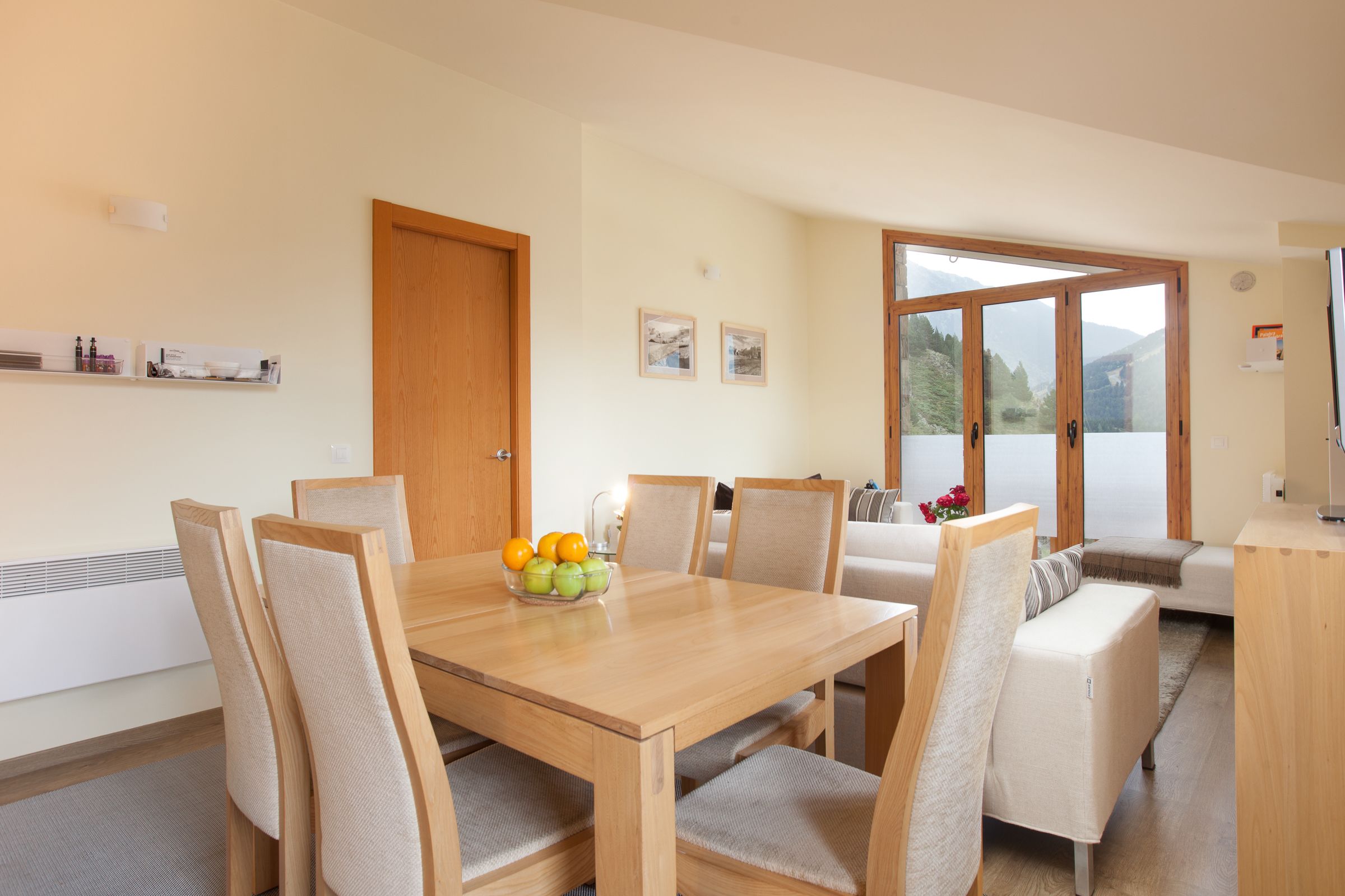 Un coin repas avec une table et des chaises en bois, un saladier rempli de fruits et une vue sur l’extérieur grâce à de grandes fenêtres.