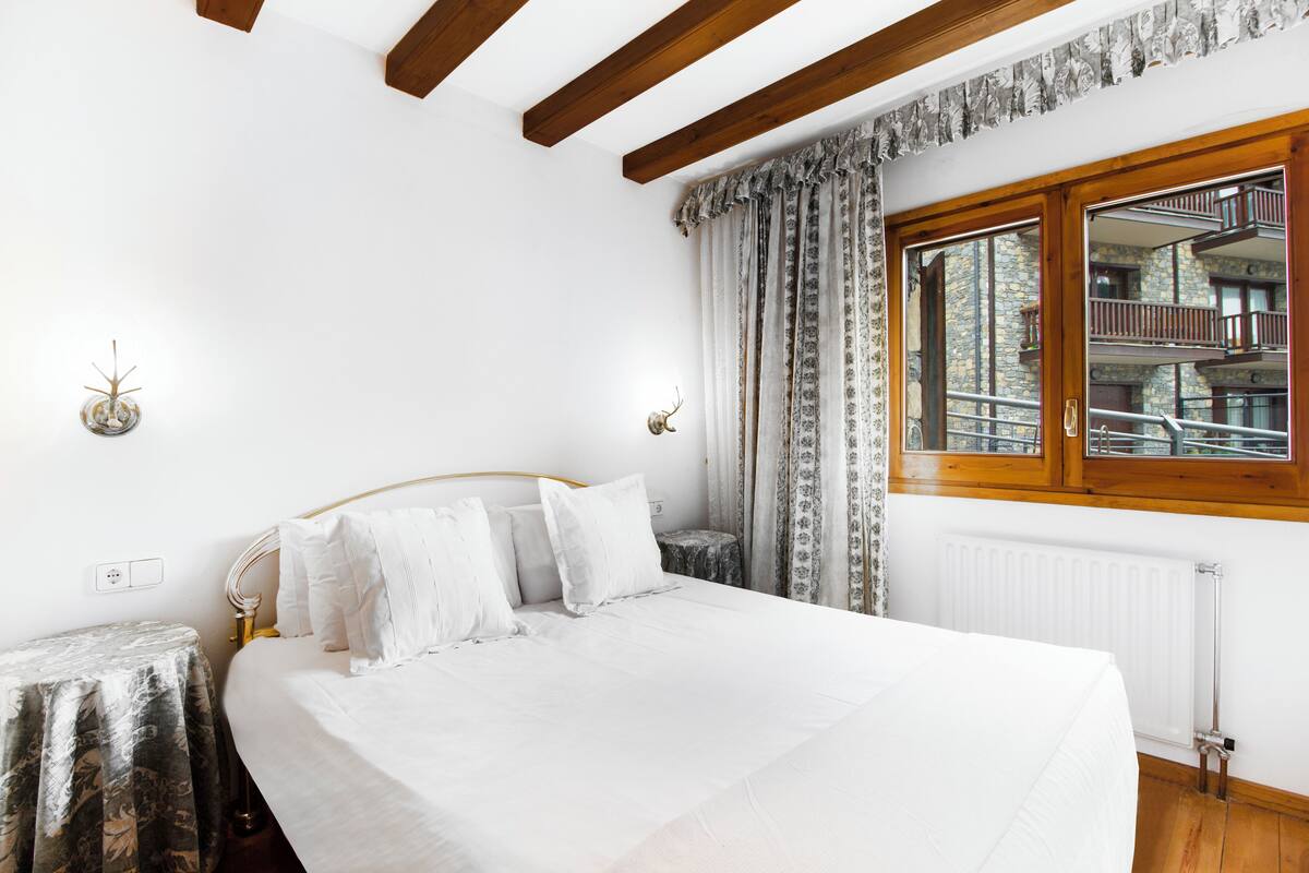 A bedroom with a large bed, wooden beams, a window with a view of a building, and a small table with a decorative item.