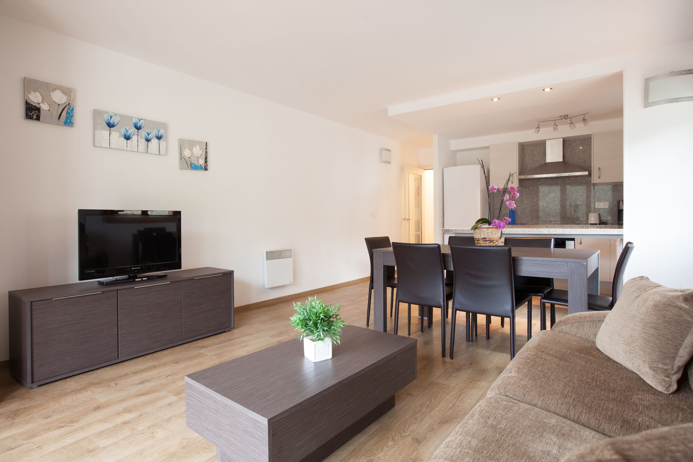 Een moderne woonkamer met een flatscreen televisie, donker houten meubels en een eethoek, met een keuken op de achtergrond.