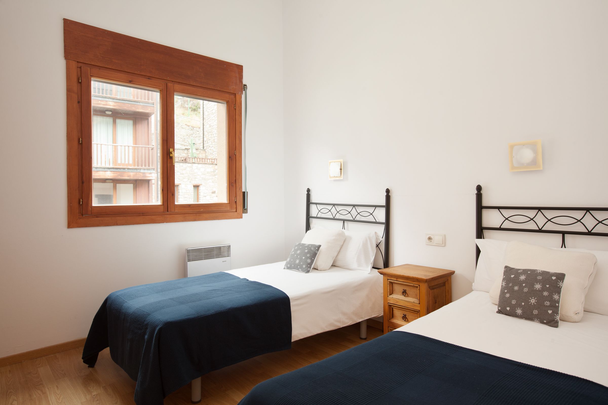 A bedroom with two single beds, a wooden nightstand, a window with a view of a balcony, and a wall-mounted light fixture.