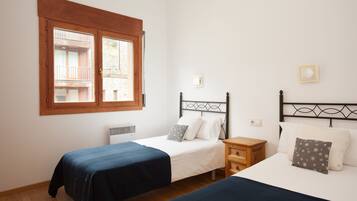 A bedroom with two single beds, a wooden nightstand, a window with a view of a balcony, and a wall-mounted light fixture.
