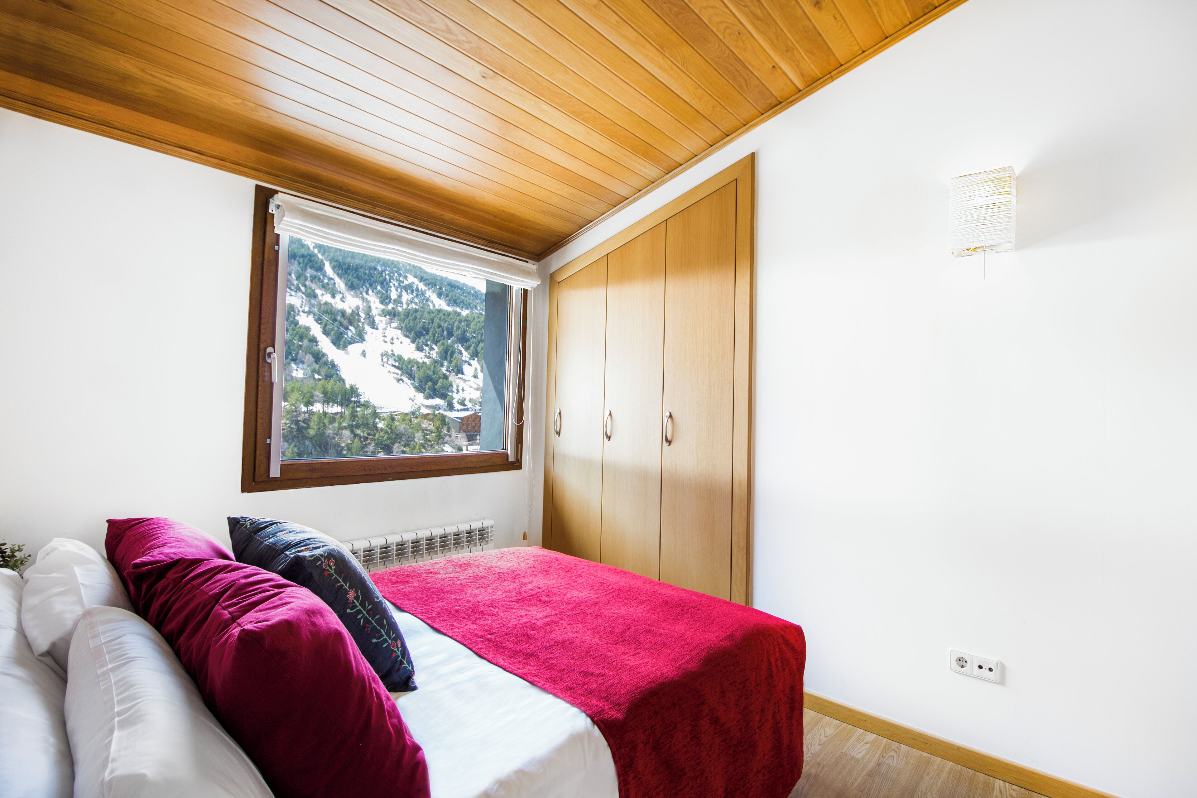 A bedroom with a bed, a window with a view of mountains, wooden wardrobe, and a radiator.