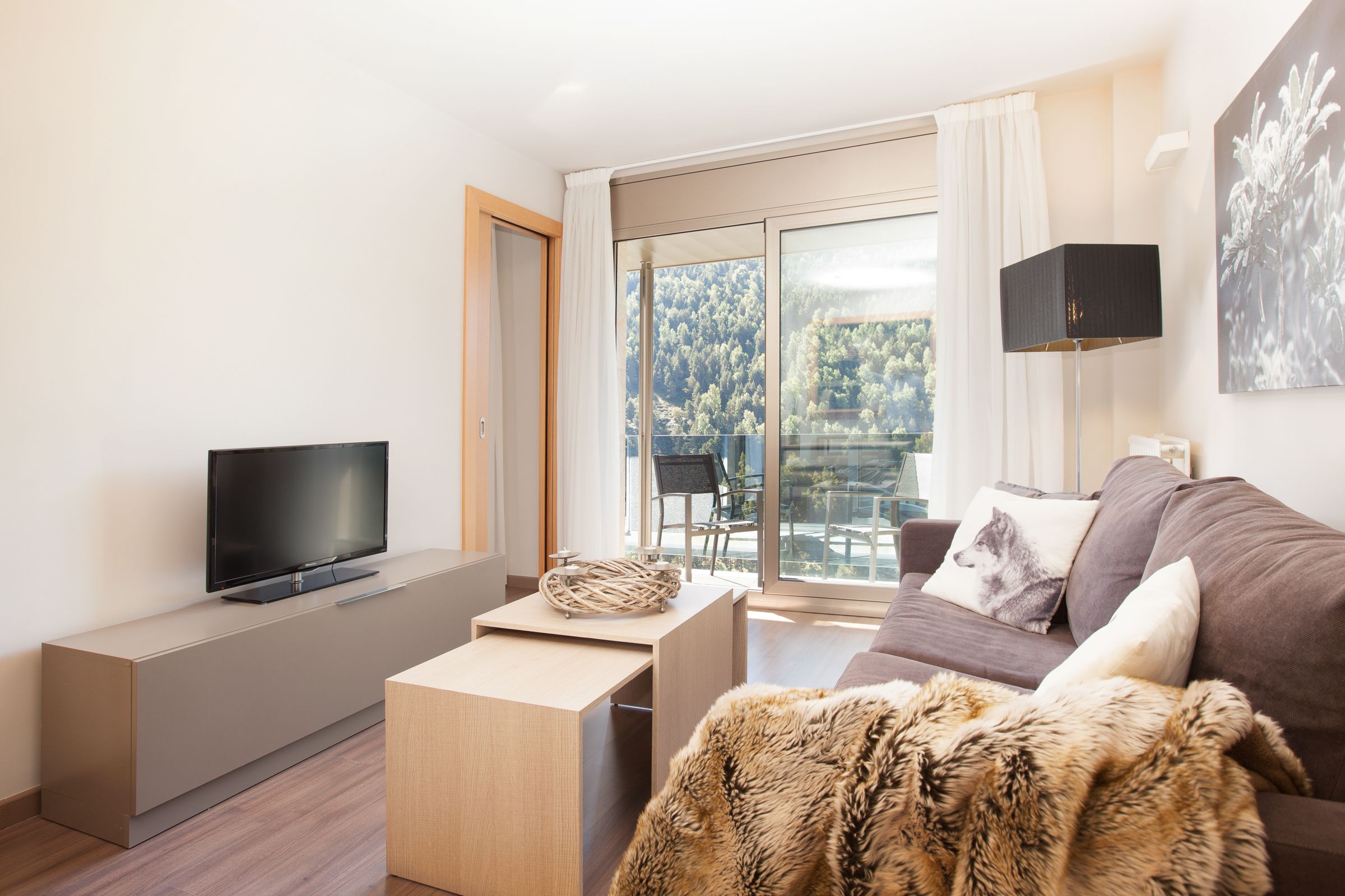 Ein modernes Wohnzimmer mit einem Flachbildfernseher auf einem schlichten Konsolentisch, einer gemütlichen Couch mit einem Fellüberwurf und einem großen Fenster mit Blick auf einen Balkon und Grünflächen.