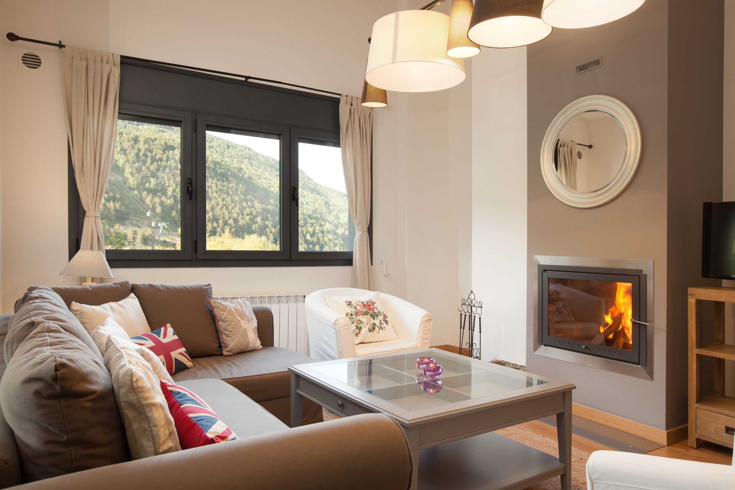 A modern living room with a sofa, armchairs, a glass coffee table, a fireplace, and a large window with curtains.