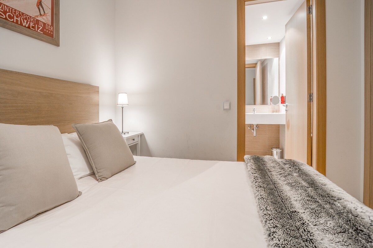 Habitación de hotel con cabecera de madera, cama con dos almohadas, mesita de noche con lámpara y baño a la vista a través de una puerta abierta.