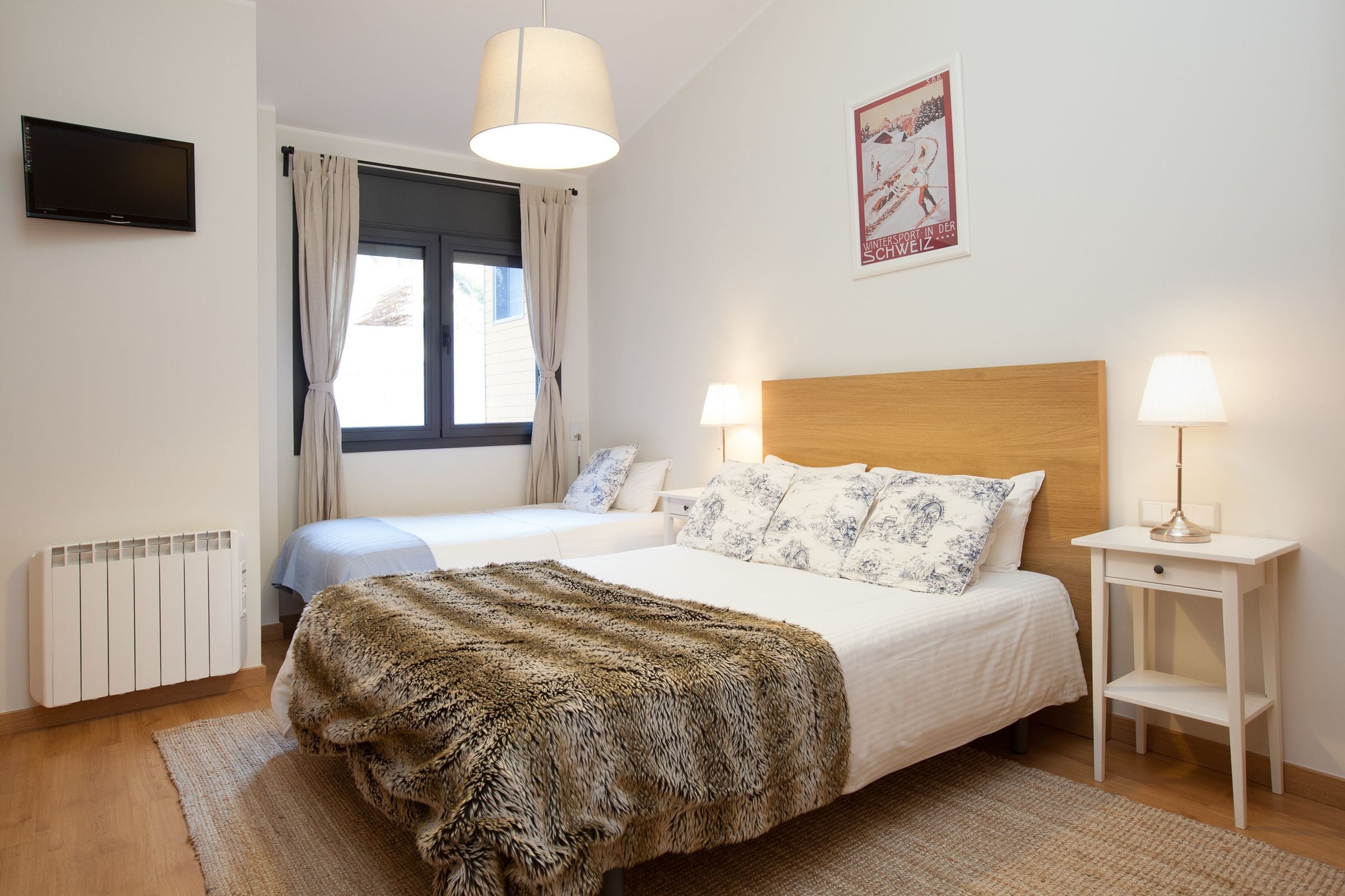 A bedroom with a large bed, two bedside tables with lamps, a radiator, a TV, and a window with curtains.