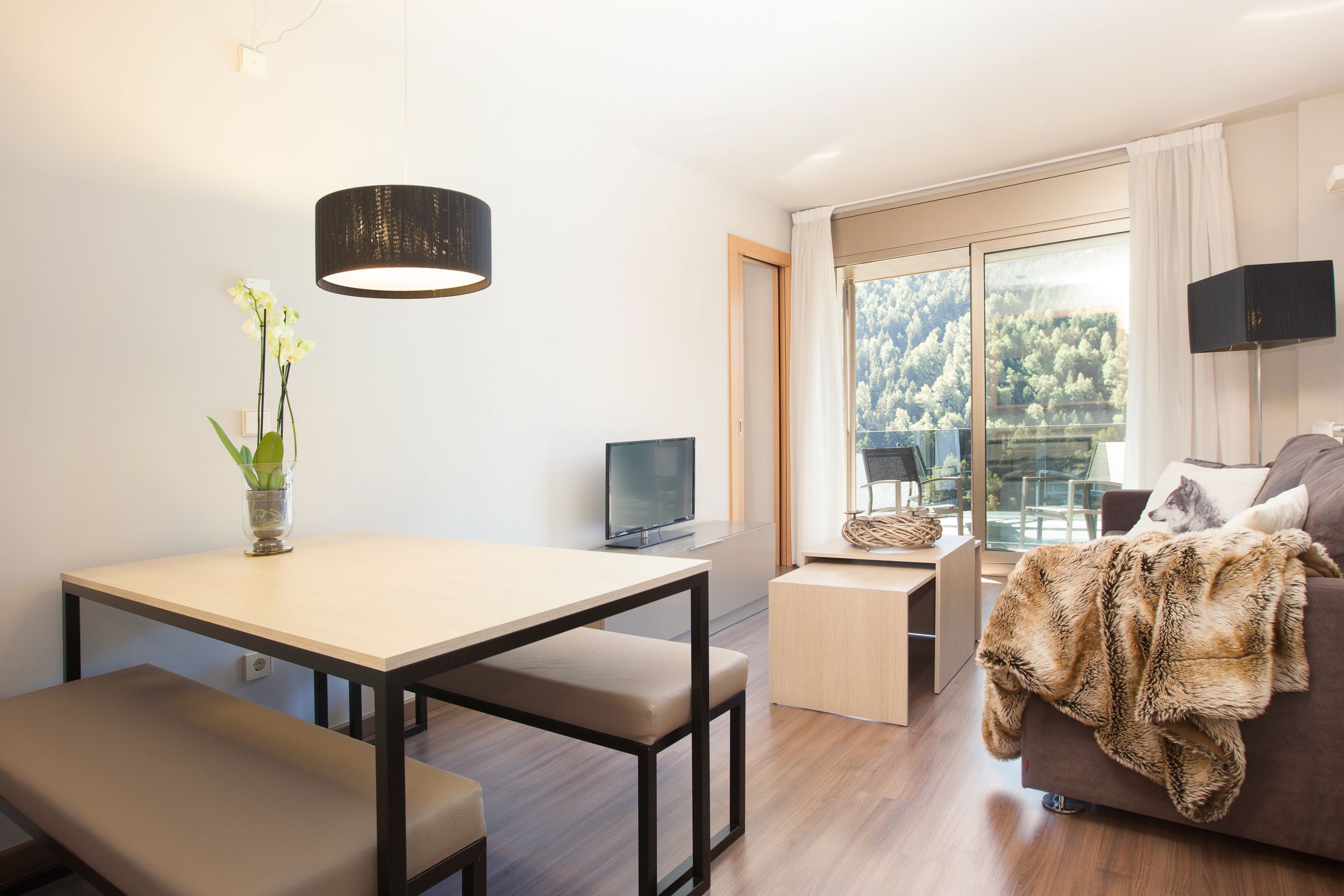 Een moderne woonkamer met een eettafel, een bankje, een bank, een televisie en een groot raam met uitzicht op bomen.