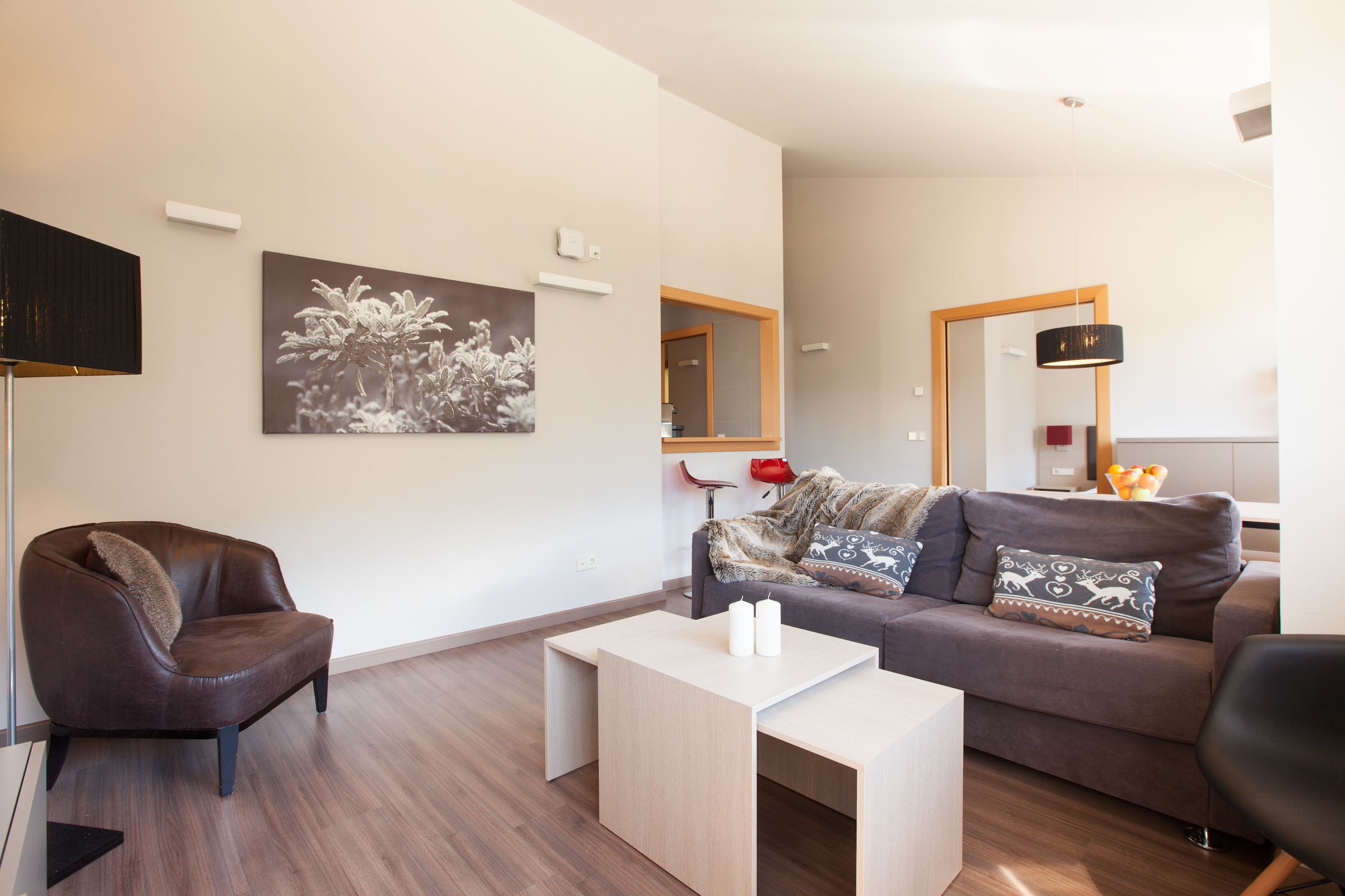 Una sala de estar moderna con un sillón de cuero marrón, un sofá gris con cojines estampados, una mesa de centro blanca y un espejo grande en la pared.