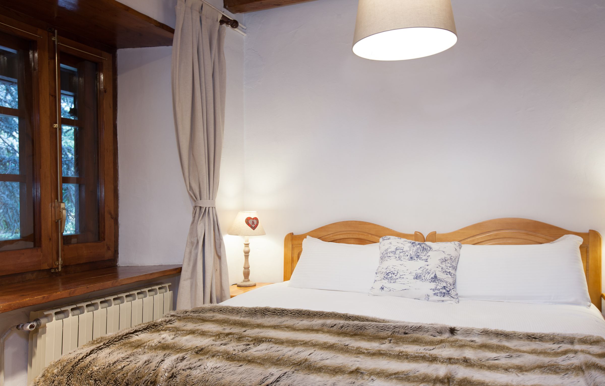 Une chambre à coucher avec une tête de lit en bois, un lit recouvert de linge de lit blanc, un plaid en fourrure, une lampe de chevet avec un abat-jour en forme de cœur et une fenêtre avec des rideaux.
