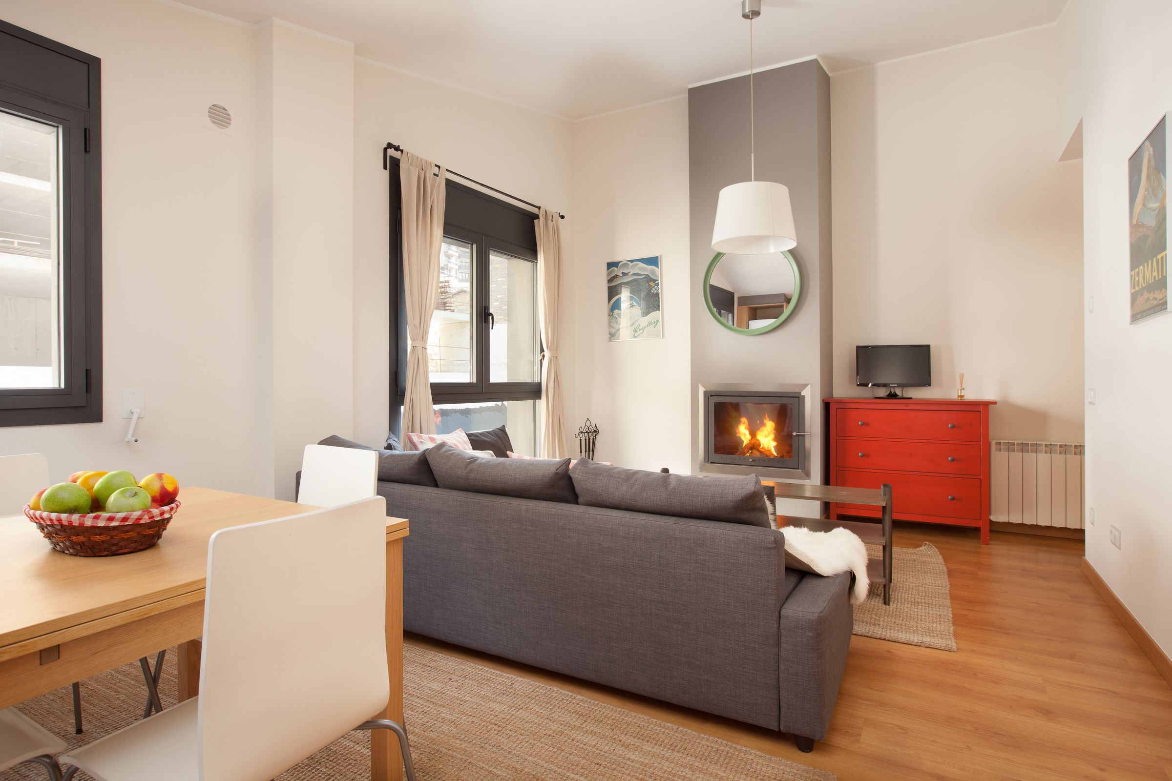 Una sala moderna con un sofá gris, una mesa de comedor de madera con una cesta de frutas, un aparador rojo, una chimenea y un televisor sobre un mueble.