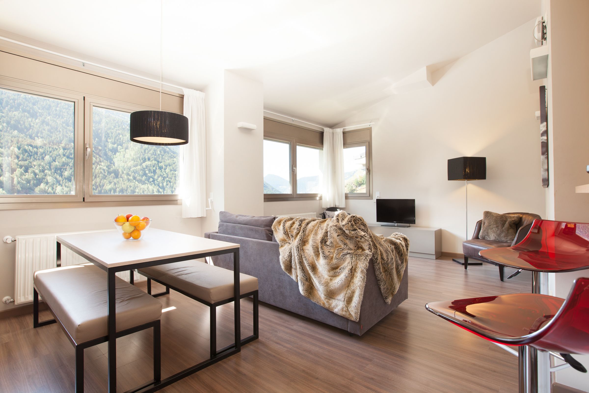 Un salon moderne comprenant un canapé, une table à manger avec un banc, un fauteuil rouge, un téléviseur et une fenêtre donnant sur des arbres.