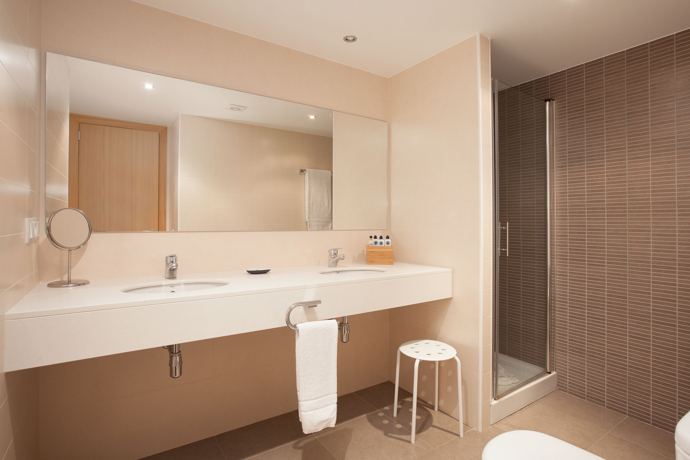 Une salle de bain moderne avec un grand miroir, deux lavabos, une douche et un petit tabouret.