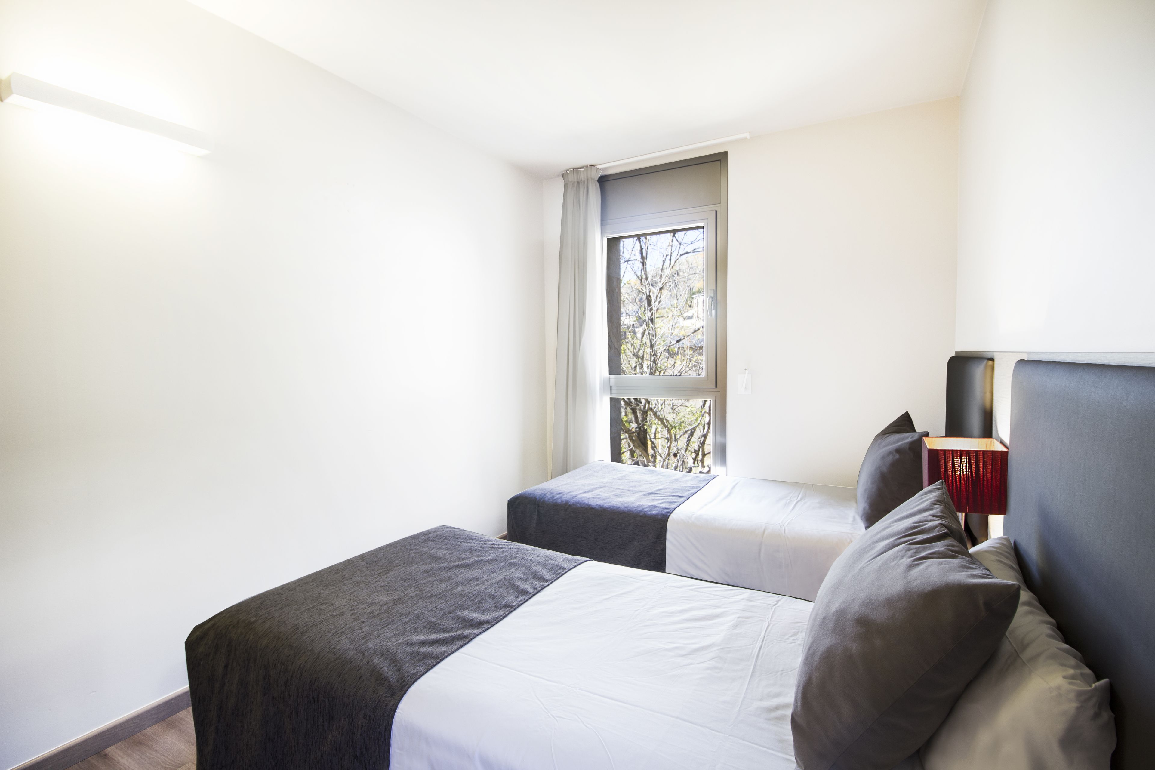 Une petite chambre moderne avec deux lits, une fenêtre donnant sur des arbres et une table de chevet avec une lampe.