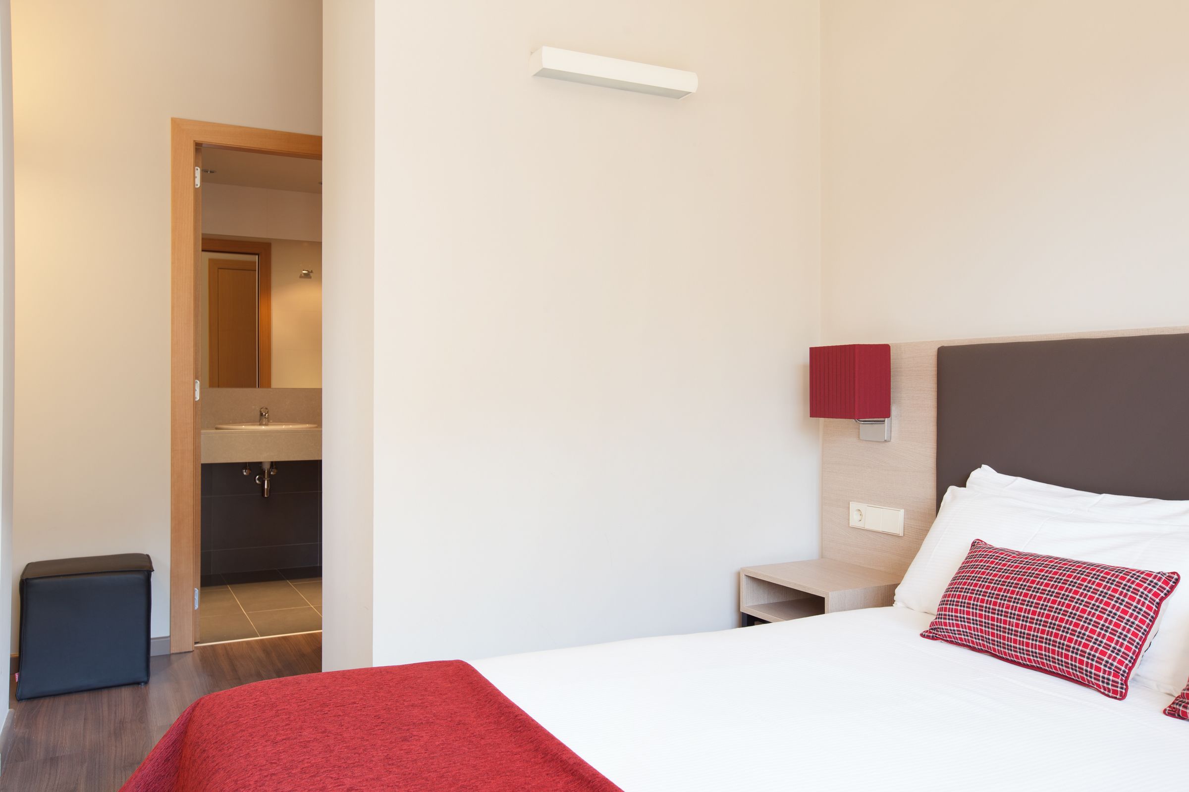 A hotel room with a bed, a nightstand, a red throw blanket, a red and black pillow, a small trash bin, and a bathroom visible through an open door.