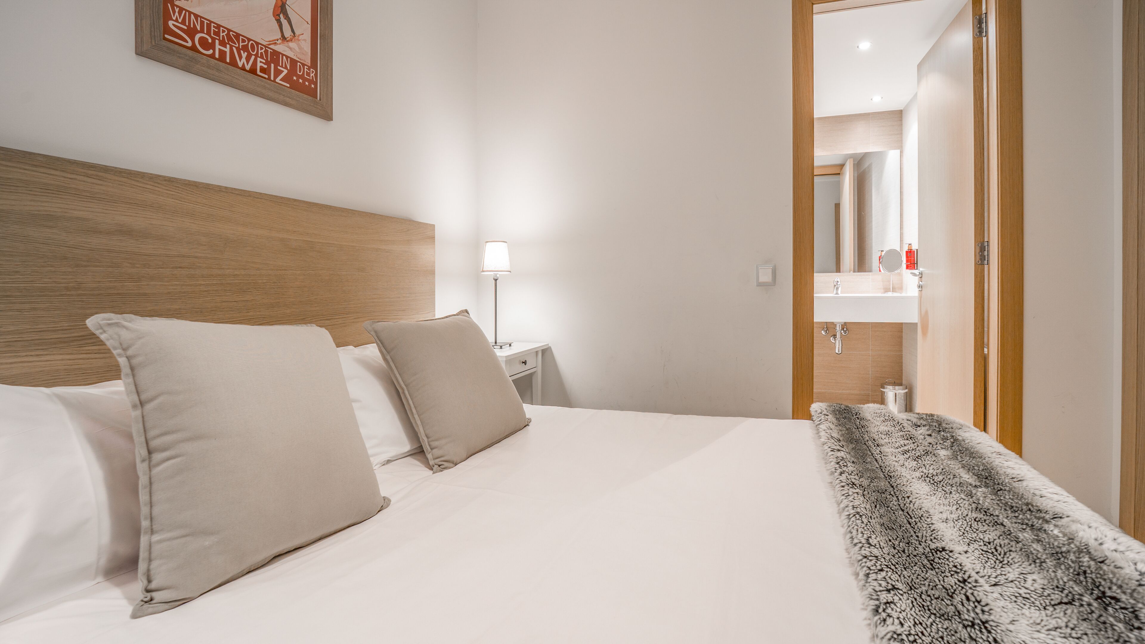 Habitación de hotel con cabecera de madera, cama con dos almohadas, mesita de noche con lámpara y baño a la vista a través de una puerta abierta.