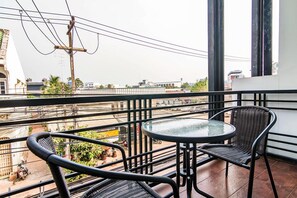 A balcony with a glass table and two wicker chairs, overlooking a street with buildings and power lines. - At Home at Udon (Udon Thani)