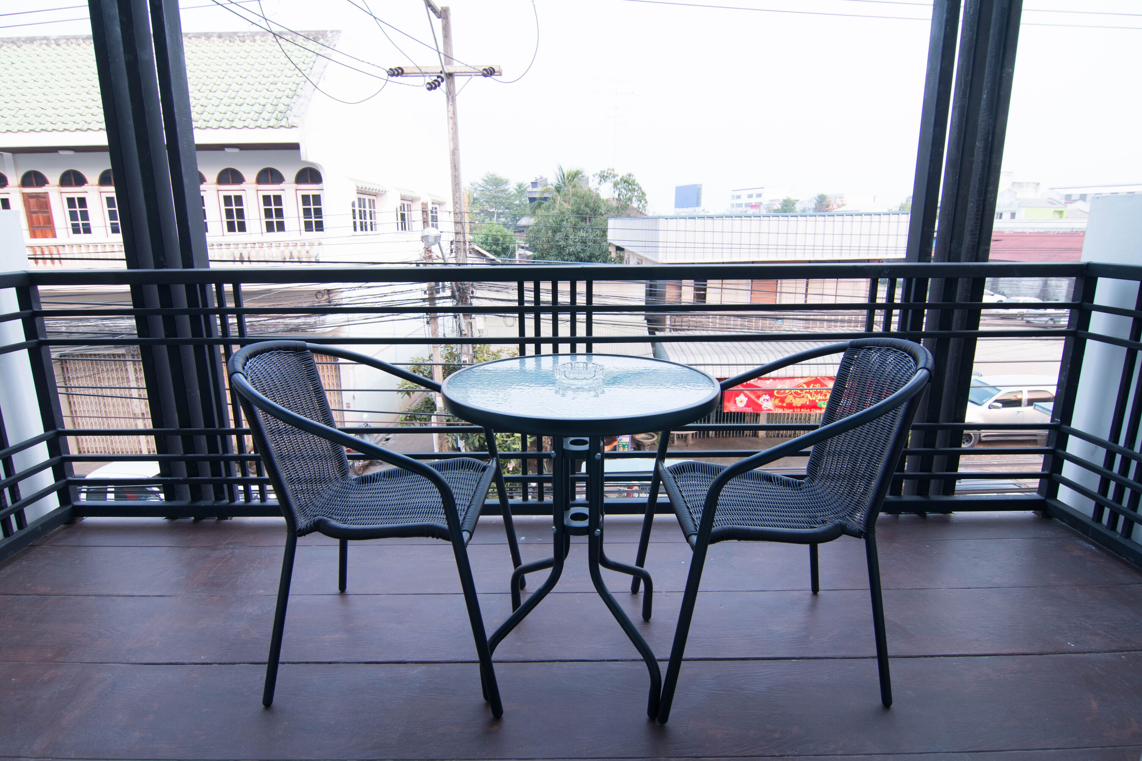 A balcony with a round table and two chairs, overlooking a street with buildings and a red vehicle.